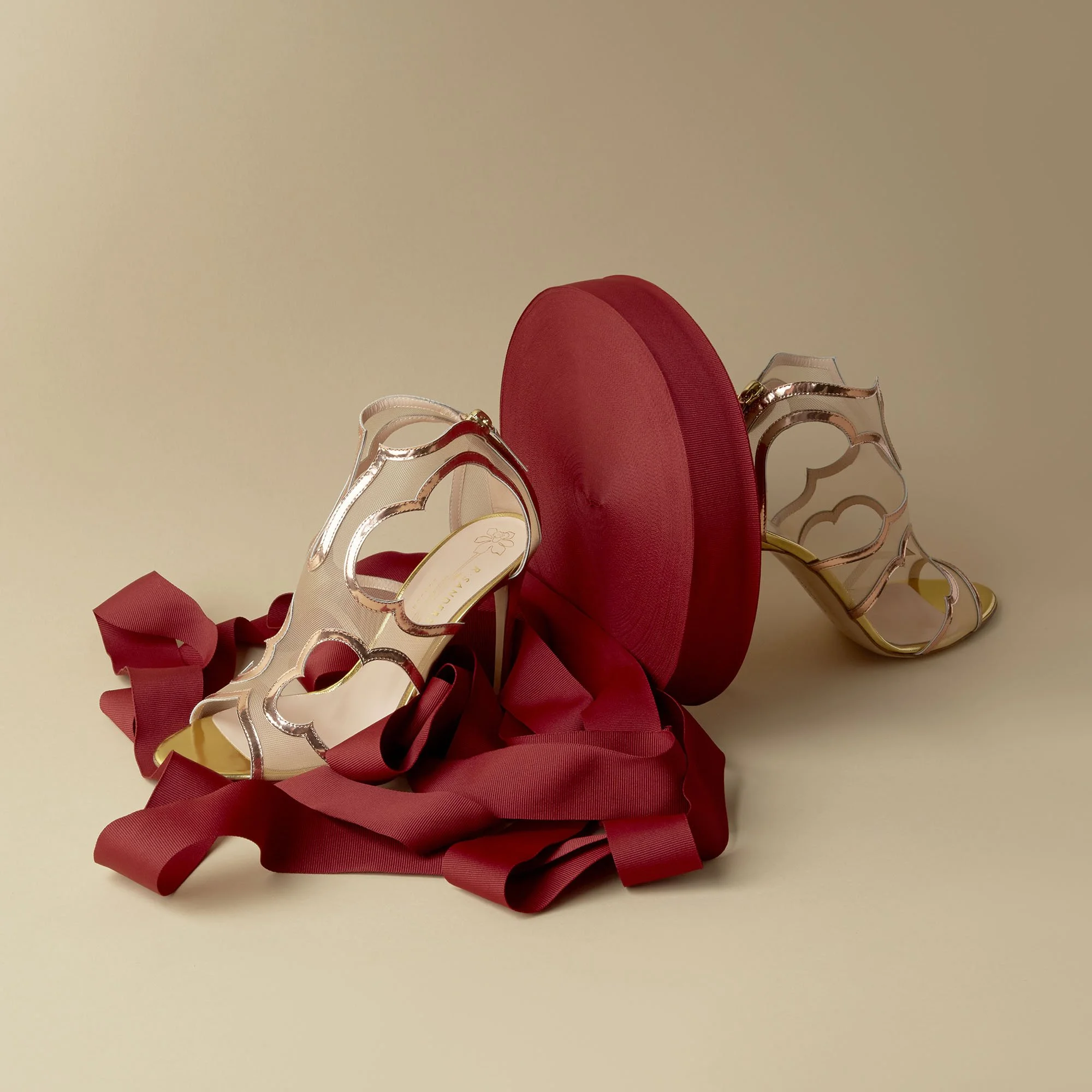A pair of clear high-heeled sandals with metallic accents, a red ribbon, and a red circular box against a beige background.