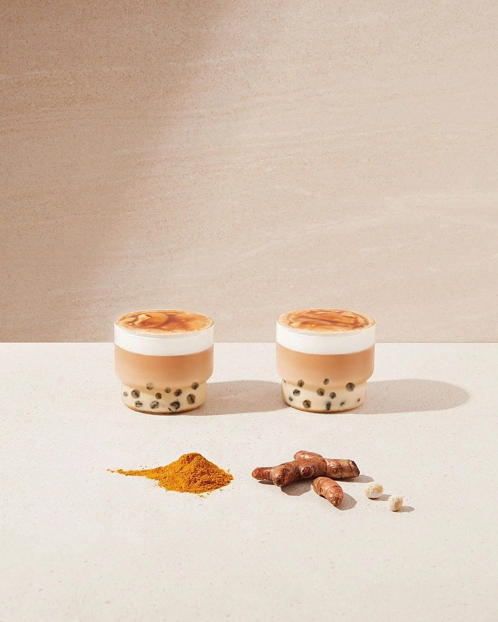 Two cups of bubble tea with tapioca pearls, turmeric powder, and fresh ginger root on a white surface against a beige background.