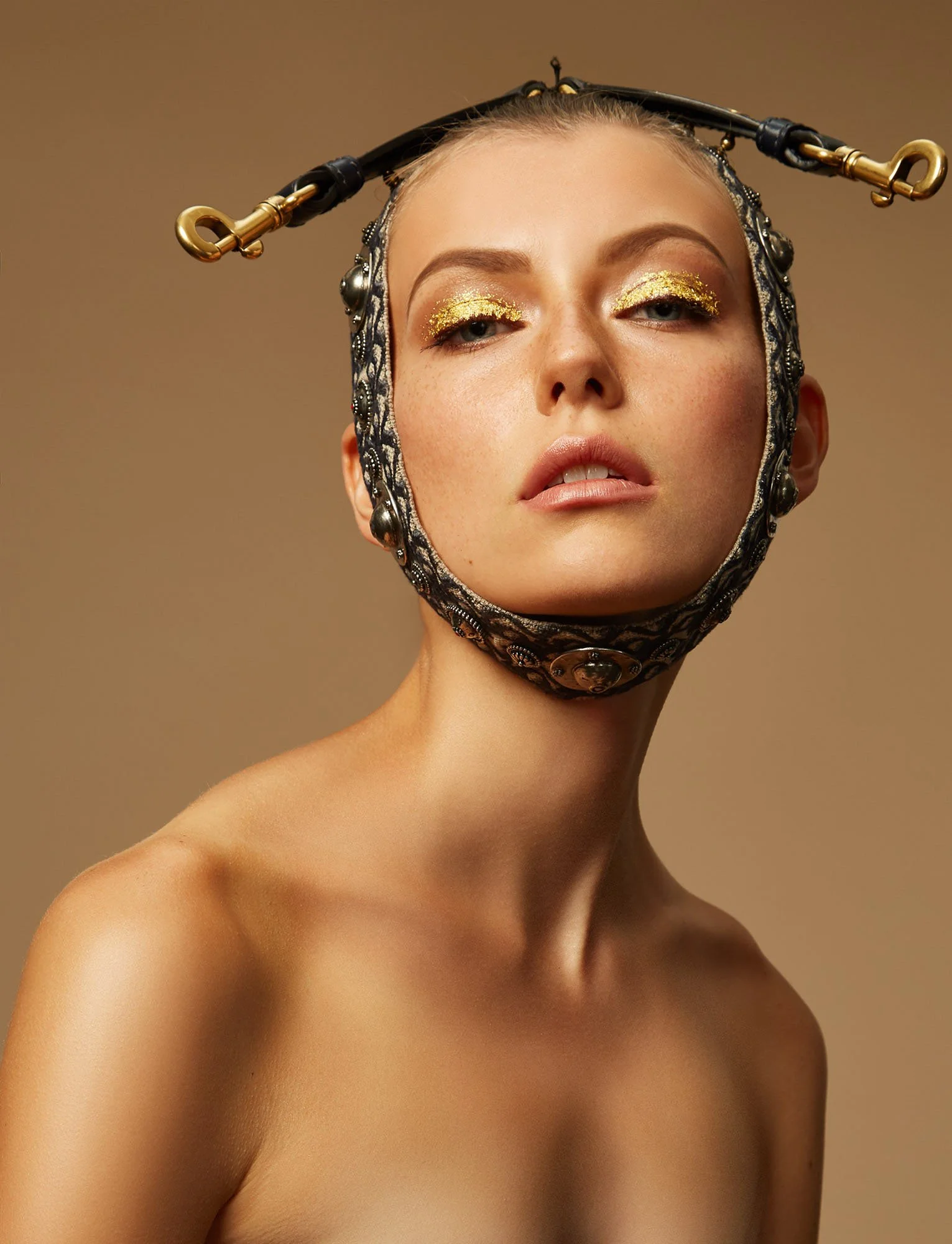 Close-up of a woman with gold eye makeup and a unique headpiece resembling a helmet with metallic and black details, against a neutral background.