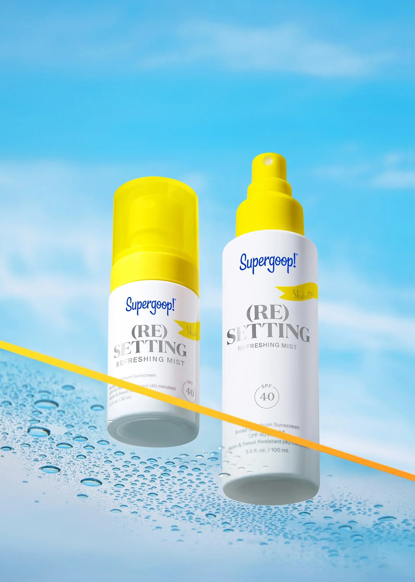 Two bottles of Supergoop! (Re) Setting Refreshing Mist sunscreen against a blue sky with clouds, with water droplets on a surface in the foreground.