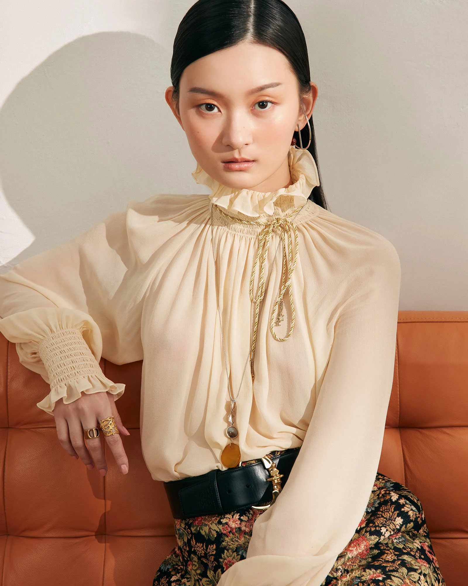 A woman with long dark hair wearing a beige ruffled high-collared blouse with a gold and beige tie, sitting on a tan leather couch, accessorized with multiple necklaces, rings, and a black belt with a gold buckle, against a plain white wall.