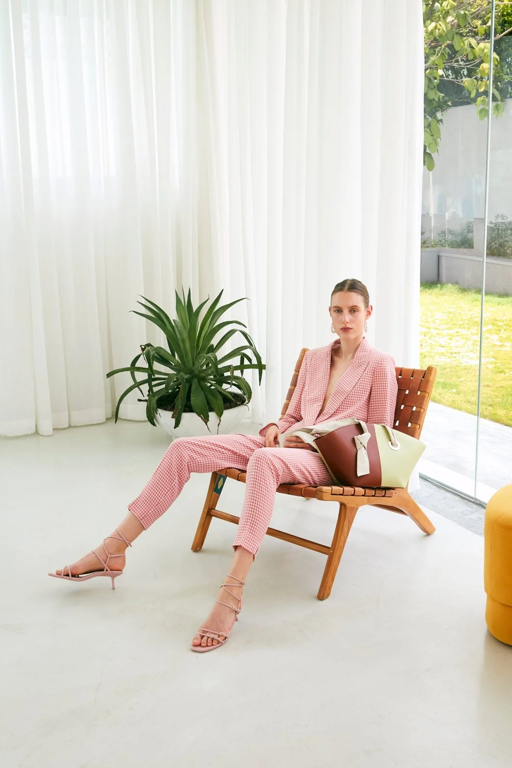 A woman in a pink checkered pantsuit with matching blazer sitting on a wooden lounge chair, holding a magazine, with a large handbag beside her, in a light-filled room with white curtains and a large green plant.