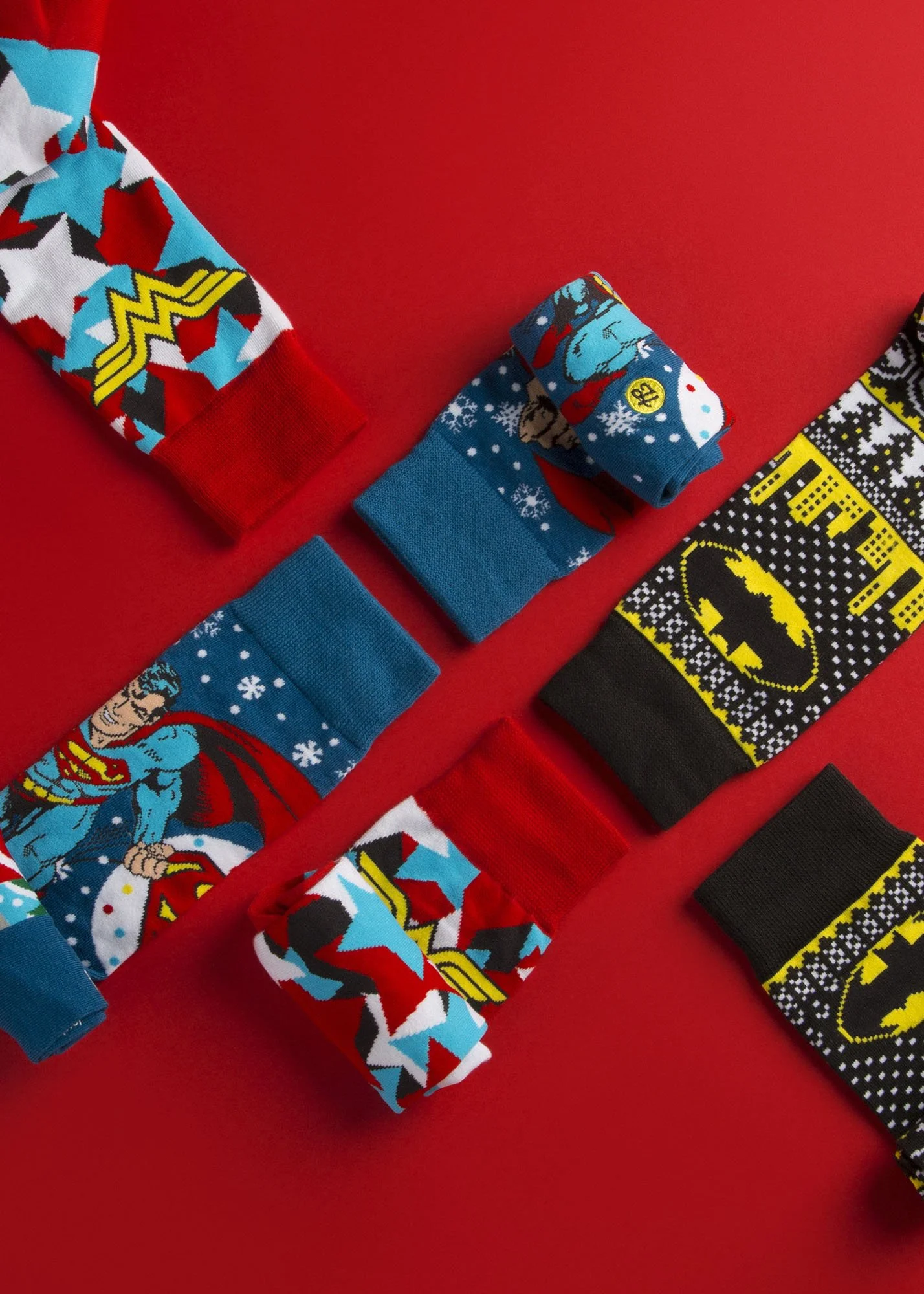 Several pairs of Christmas-themed socks with superhero and comic book patterns, including Wonder Woman and Batman, are laid out on a red surface.