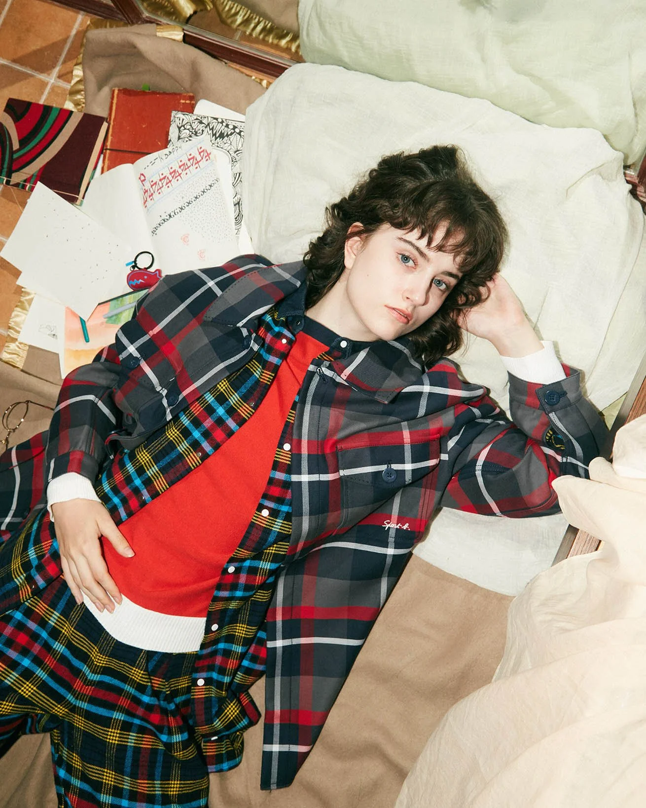 A woman with curly brown hair lying on a bed, wearing a red shirt under a plaid blazer, plaid pants, and surrounded by papers and books.