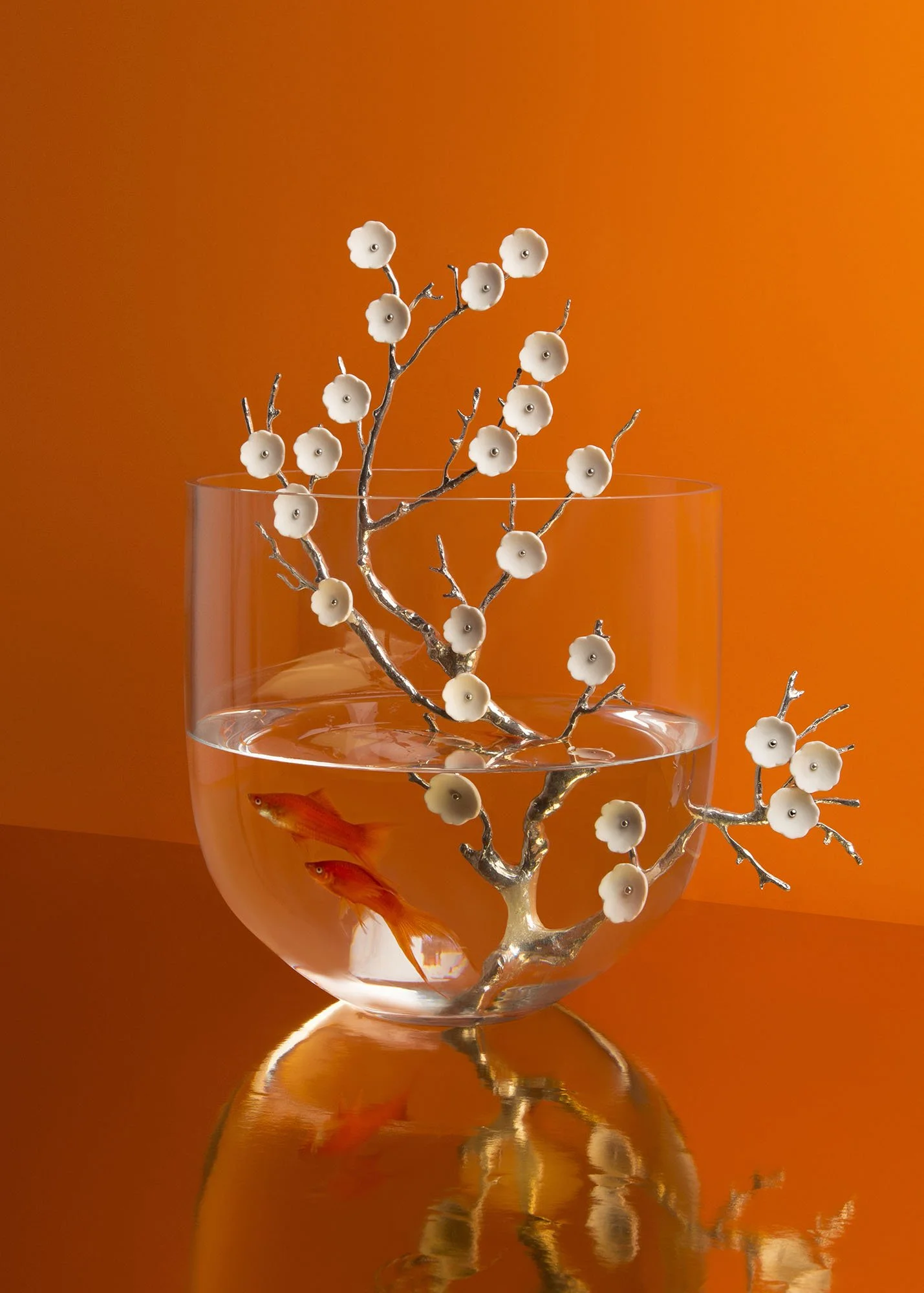 A transparent glass bowl with water and two goldfish inside, holding decorative silver branches with white flower-shaped ornaments, set against an orange background.
