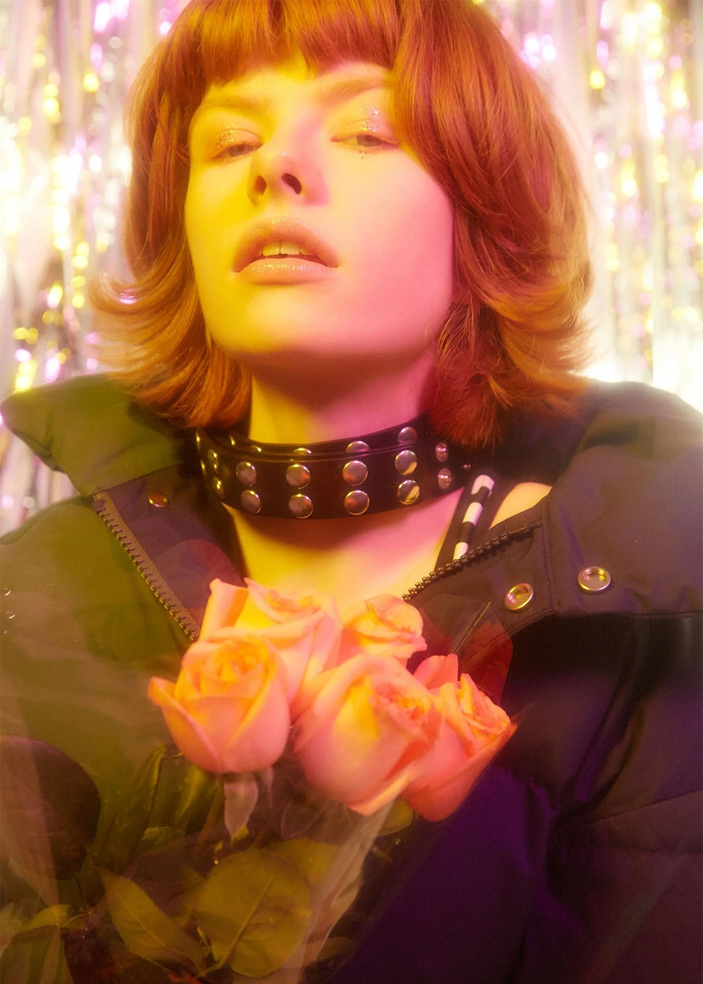A woman with shoulder-length reddish hair, wearing a black spiked choker and black jacket, holding a bouquet of yellow roses, with colorful glittery background.