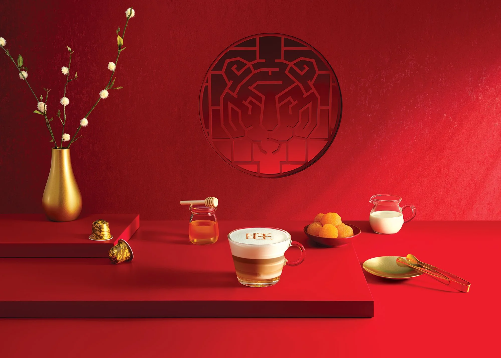 Red table setting with a gold vase holding white flowers, a jar of honey, a coffee cup with latte art, a bowl of orange fruit, a small pitcher of milk, and gold utensils, with a red wall and a lion emblem in the background.