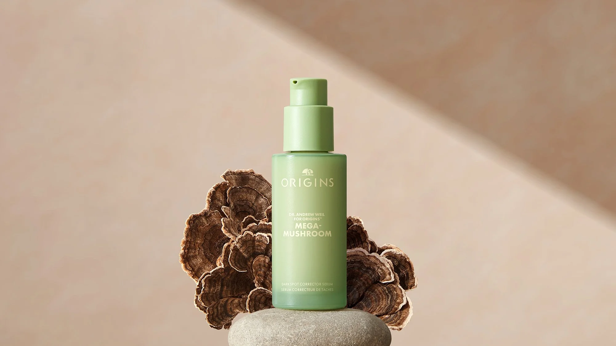 A green bottle of Origins Mega-Mushroom Darkspot Corrector Serum on a stone with mushroom-shaped wood slices behind it.