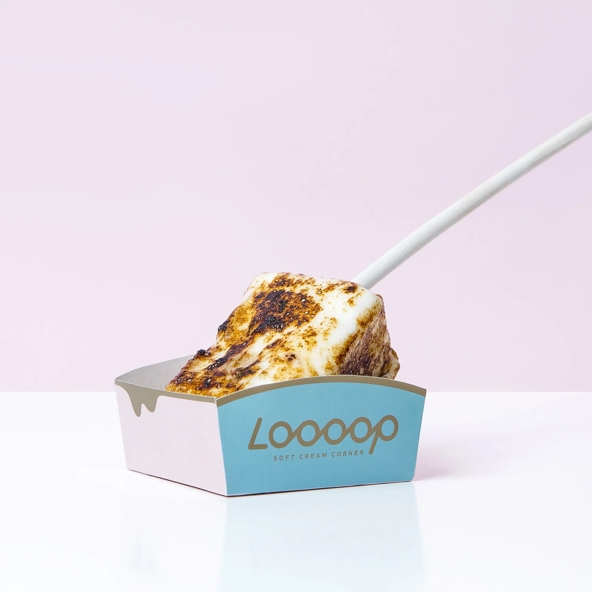 A s'mores marshmallow with a golden brown, slightly burnt surface in a blue and white paper container labeled 'Loooop Soft Cream Corner' on a white surface against a light pink background.