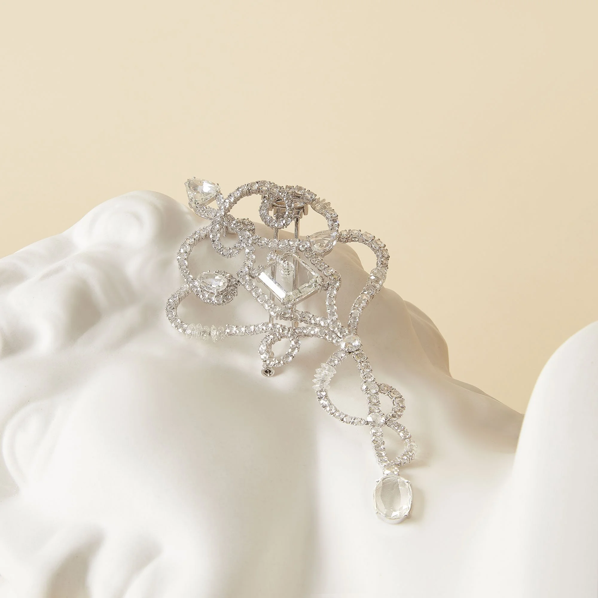 A silver, diamond-encrusted brooch with a floral and swirl design, featuring a dangling clear gemstone, resting on a white ceramic surface against a beige background.