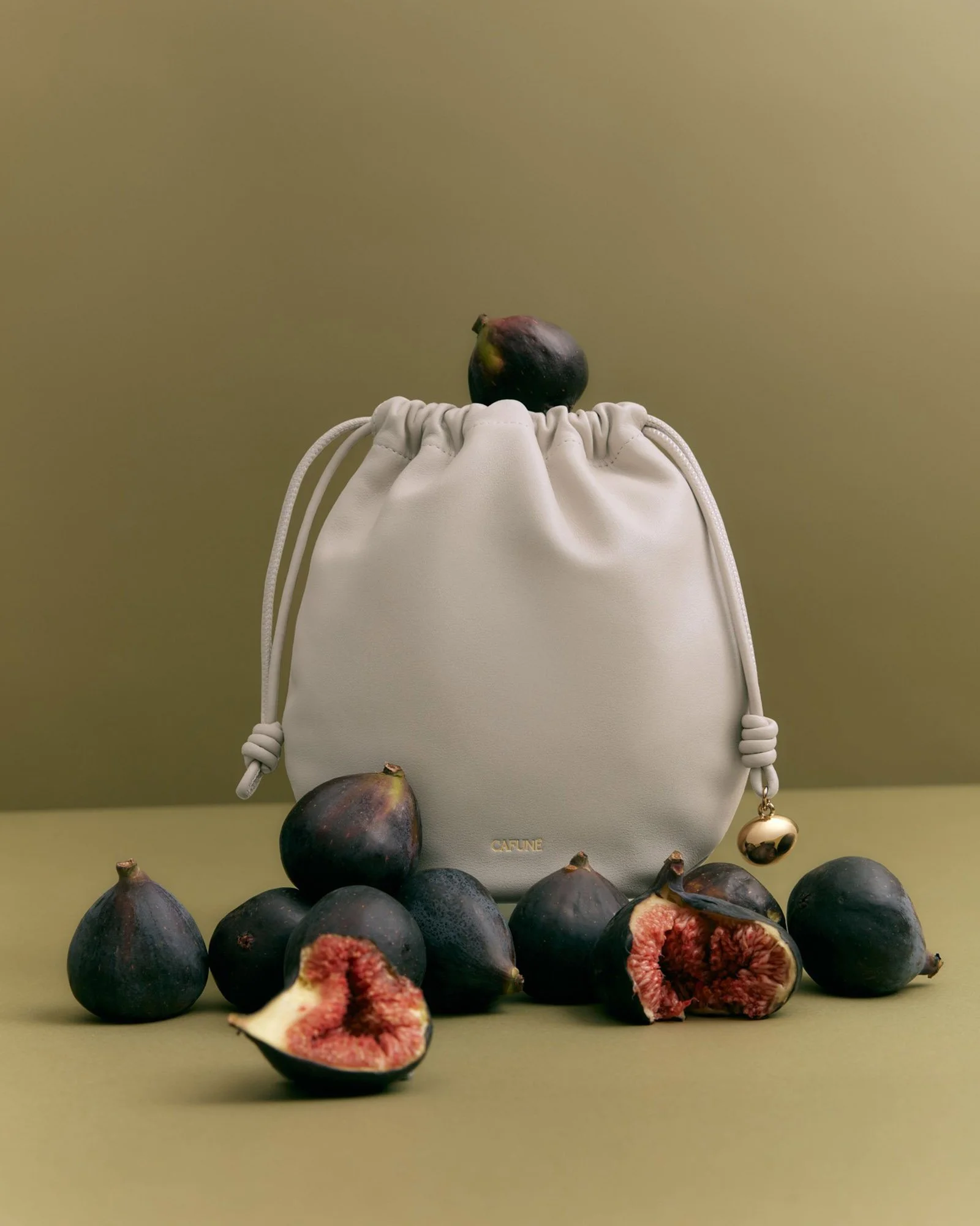A white designer bag with the brand name 'CAFEINE' embossed in gold letters, surrounded by whole and sliced figs on a green background.
