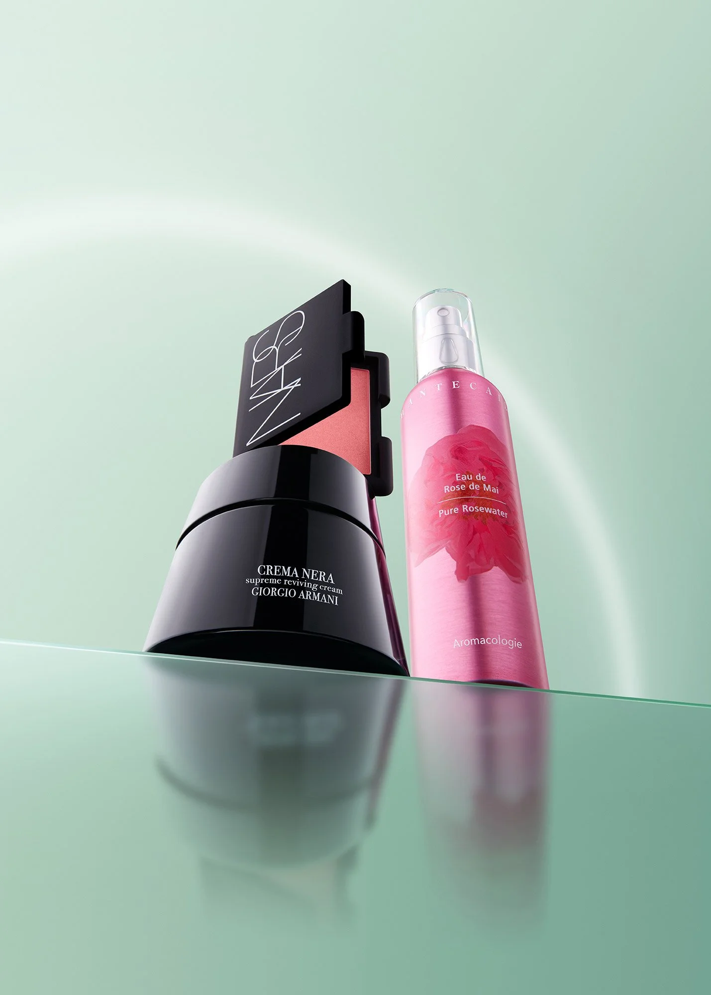 A black container labeled Crema Nera Supreme Reviving Cream from Giorgio Armani, with a pink makeup blush or compact, and a pink spray bottle of Eau de Rose de Mai, Pure Rosewater, from Inta.
