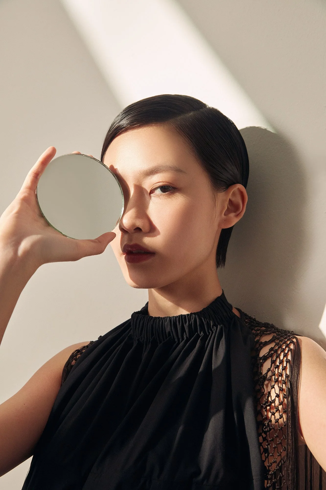A woman with short dark hair holding a round mirror/image up to her face while leaning against a white wall, with sunlight creating shadows.
