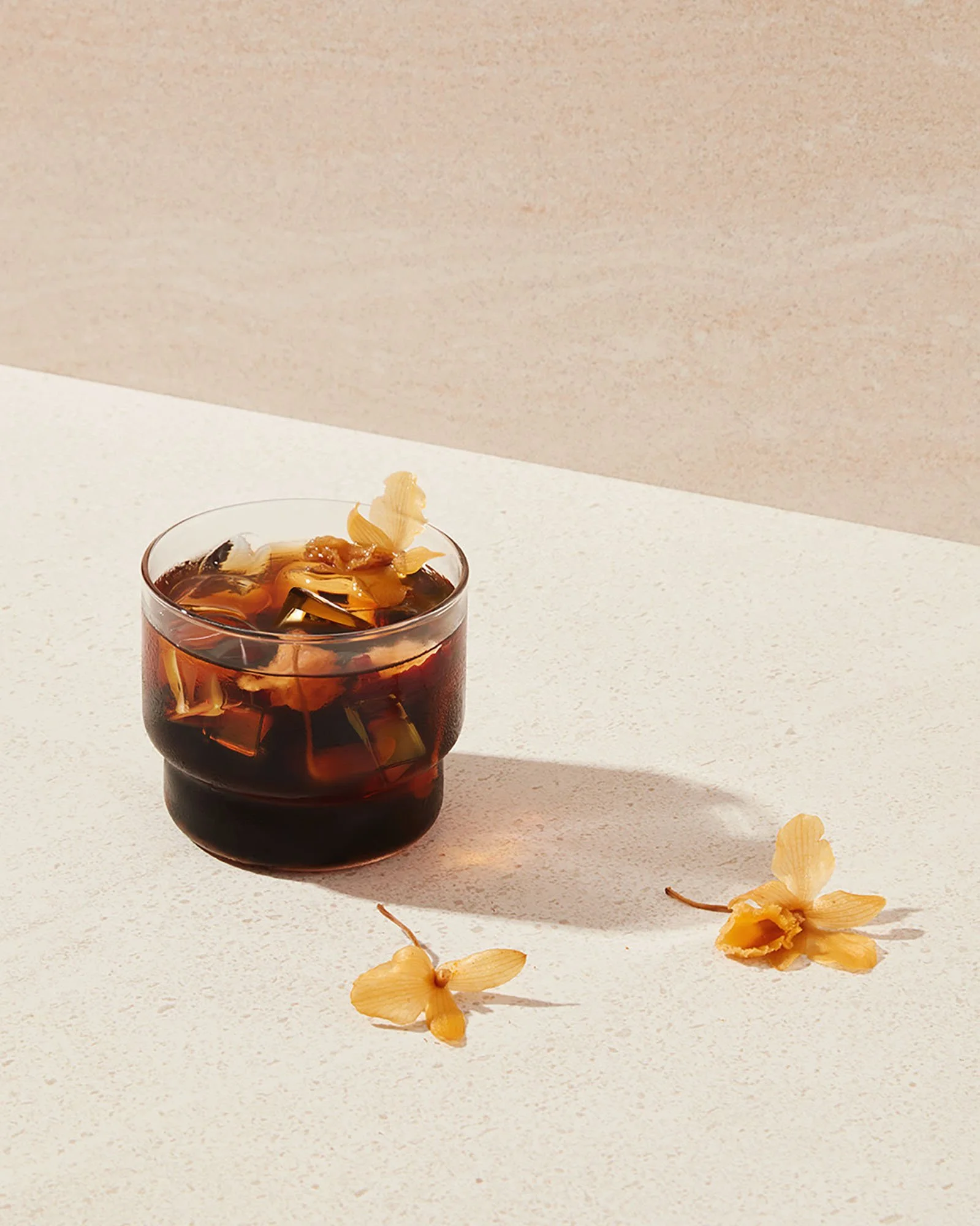 A glass of iced coffee with ice cubes and flower-shaped decorations on top, placed on a white surface with two additional flowers nearby, casting a shadow.