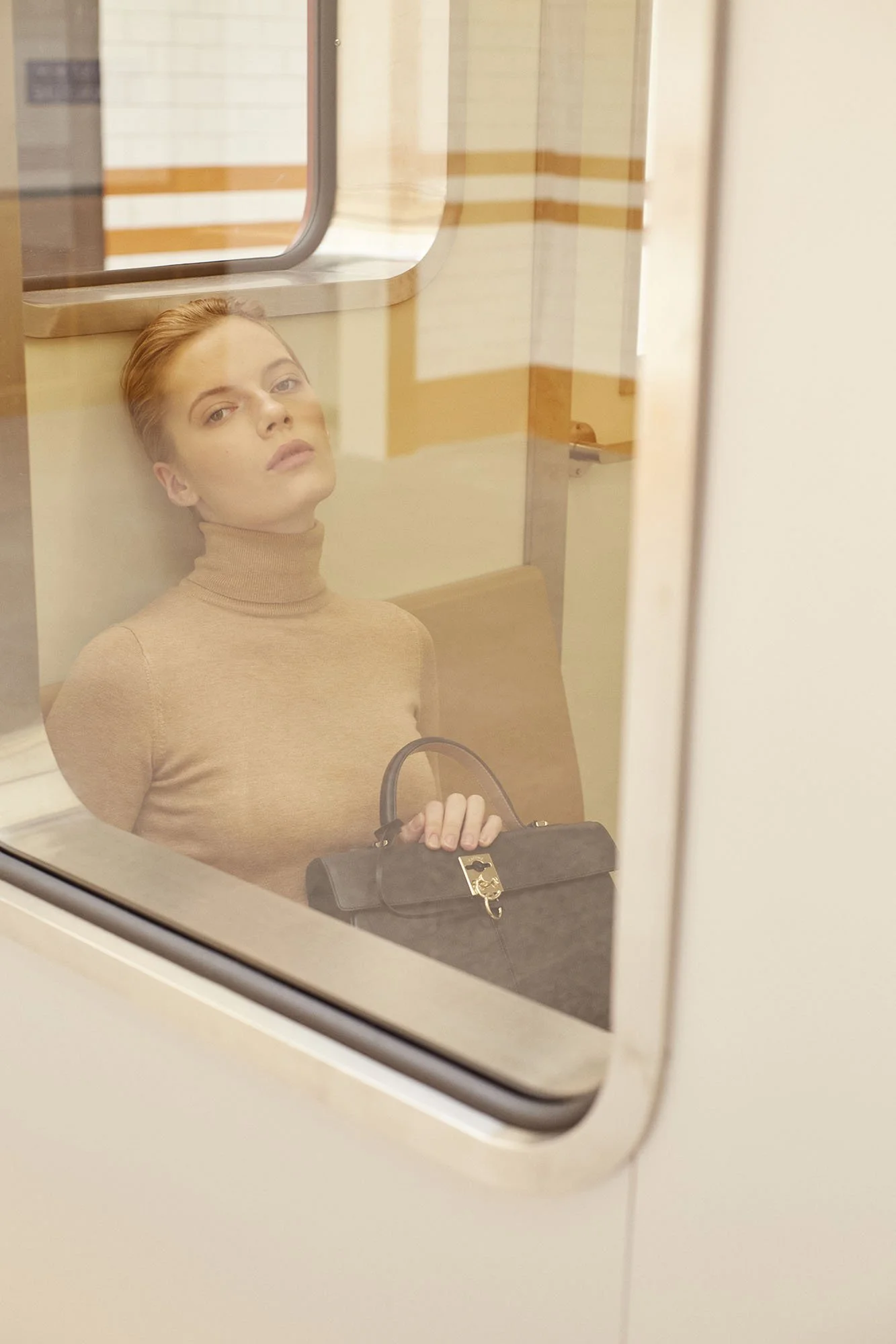 A woman with red hair and a beige turtleneck sweater sitting on a bench in a room, viewed through a glass window or door, holding a black handbag on her lap.