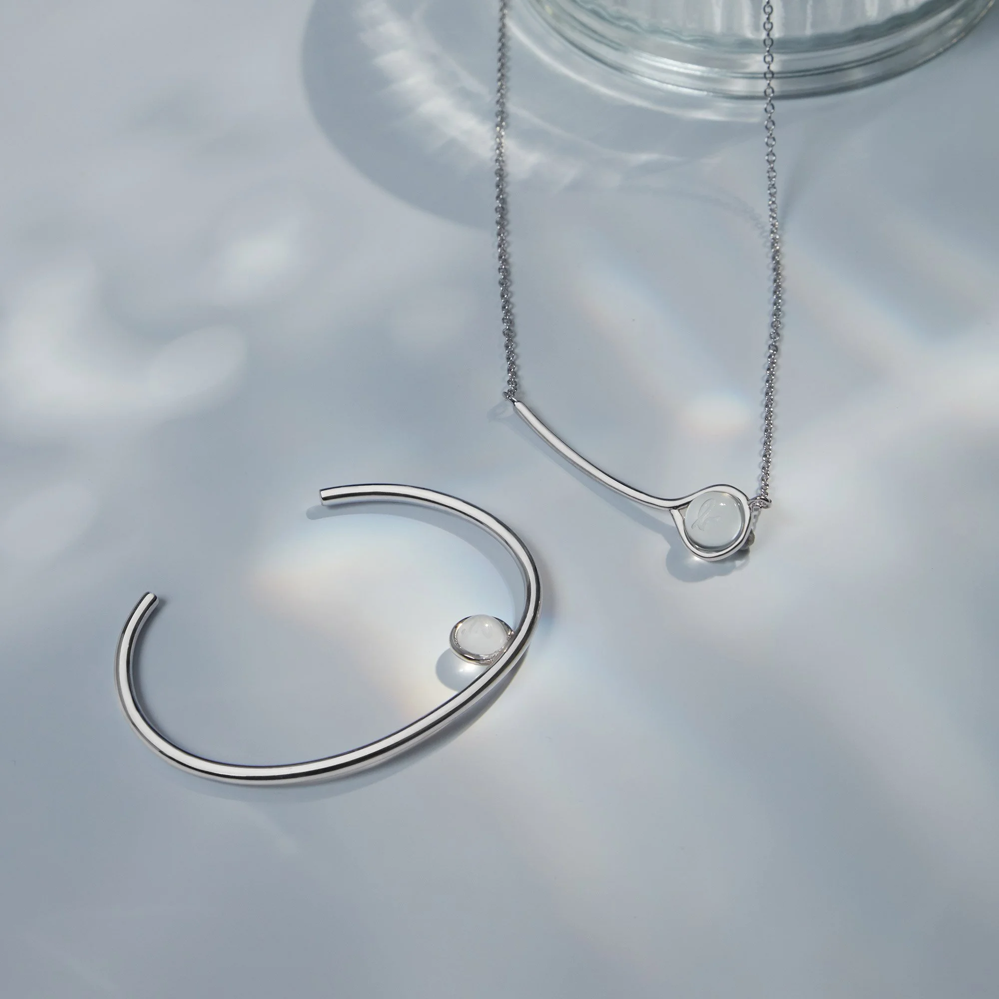 Silver jewelry set with a necklace and bangle, each featuring a small round pendant, on a reflective surface with light reflections.