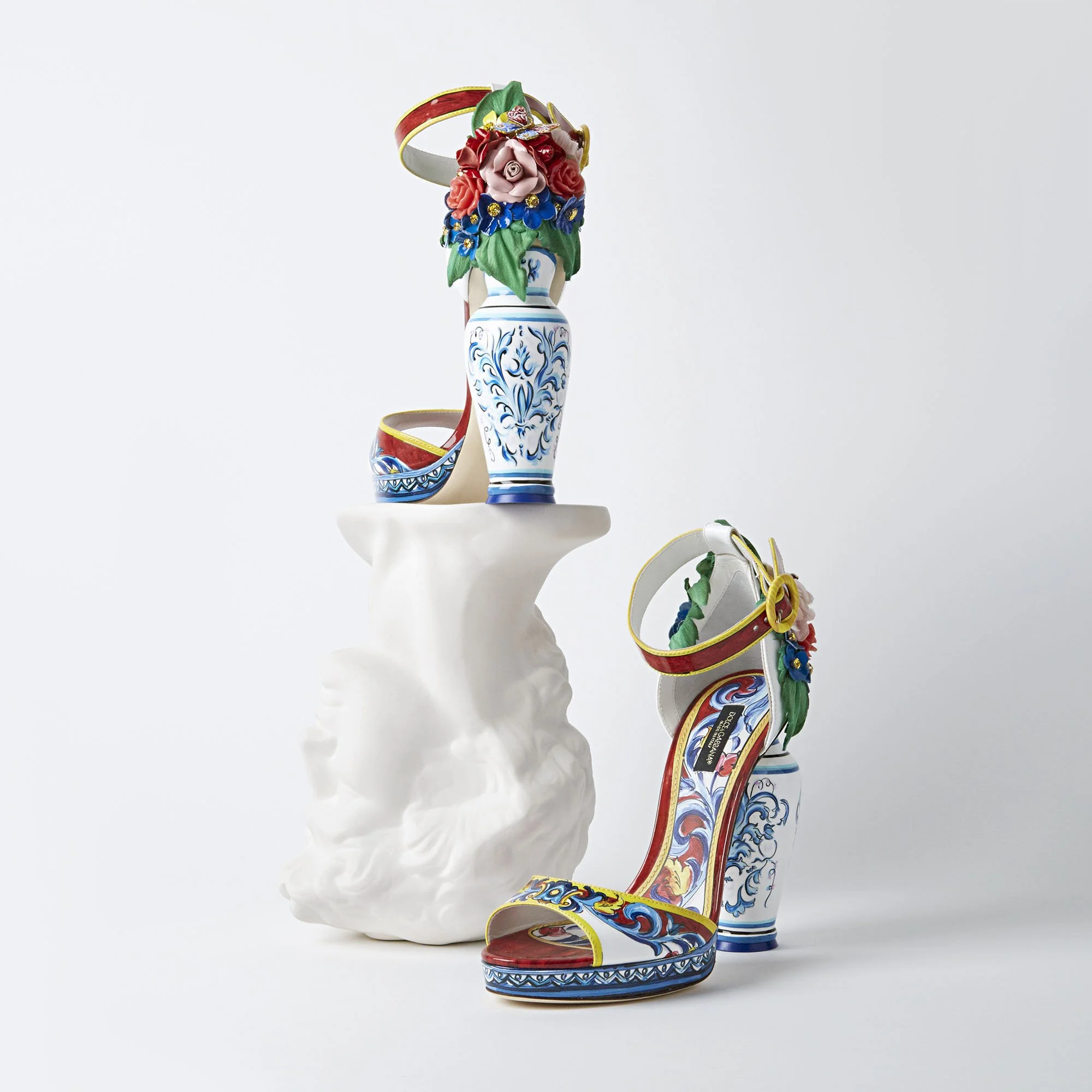 Colorful high-heeled shoes with decorative floral and stripe designs, placed on a white abstract pedestal with a plain white background.