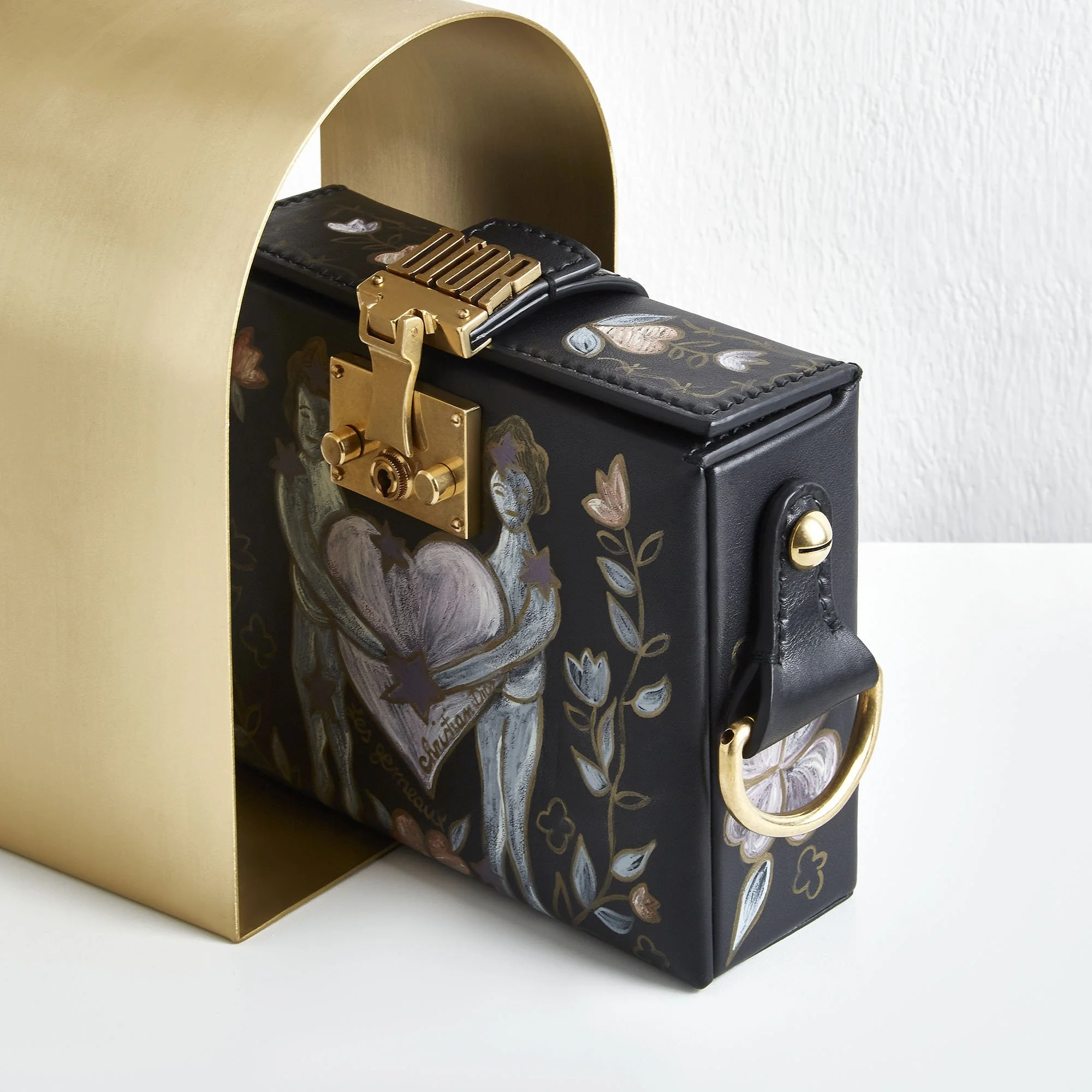 A black handbag with artistic painted illustrations, gold hardware, and a gold handle, placed inside a gold-colored box.
