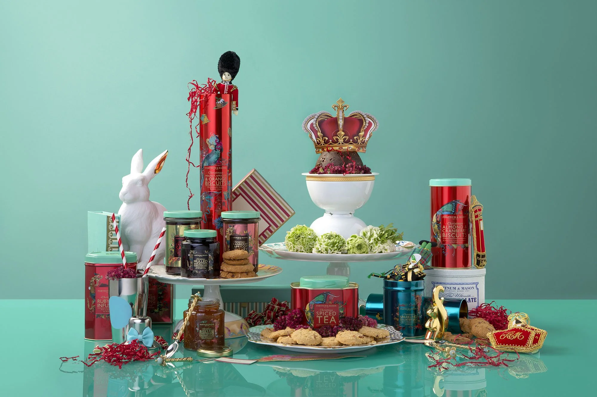 Assorted Christmas-themed treats and decorations on a teal table, including cookies, jam, biscuits, a white rabbit figurine, a toy soldier, a crown, and holiday tins.