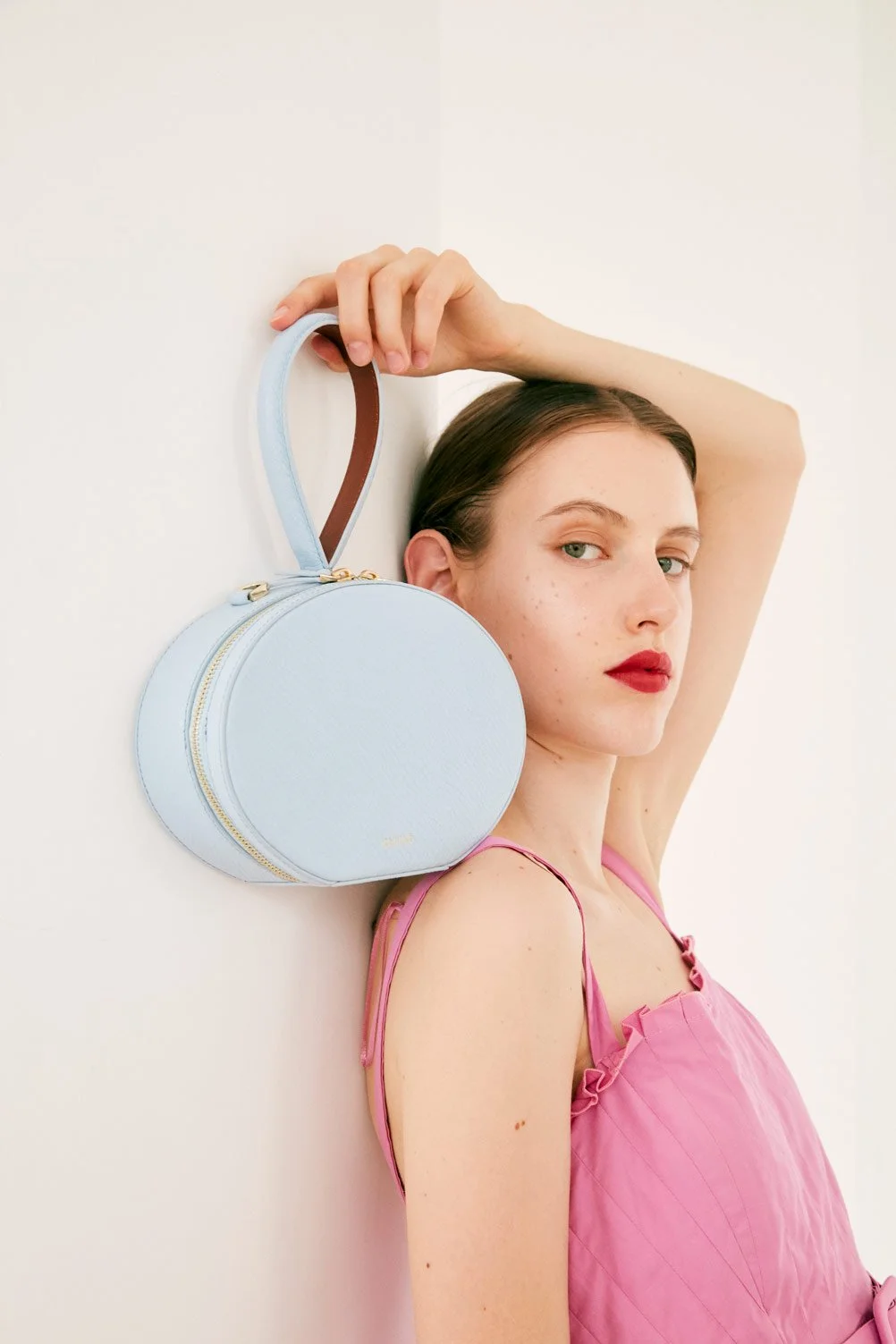 A woman with red lipstick and freckles, wearing a pink top, leans against a white wall with her arm raised above her head, holding a round, light blue handbag with a brown handle.