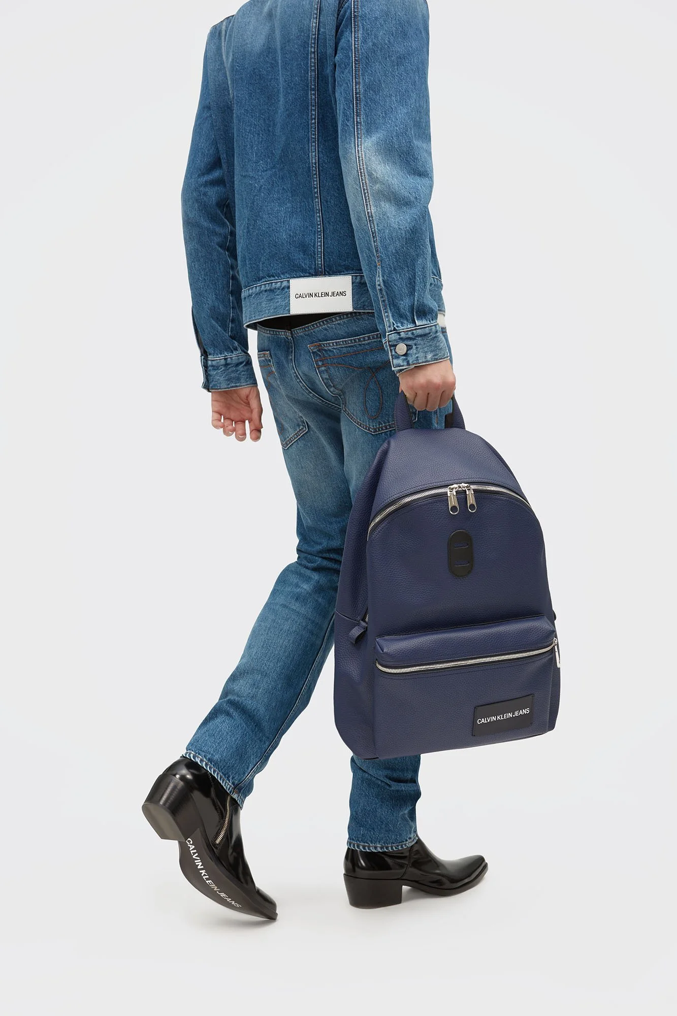 Person dressed in blue denim jacket and jeans, holding a navy backpack, standing on black shiny boots with white text, against a plain white background.