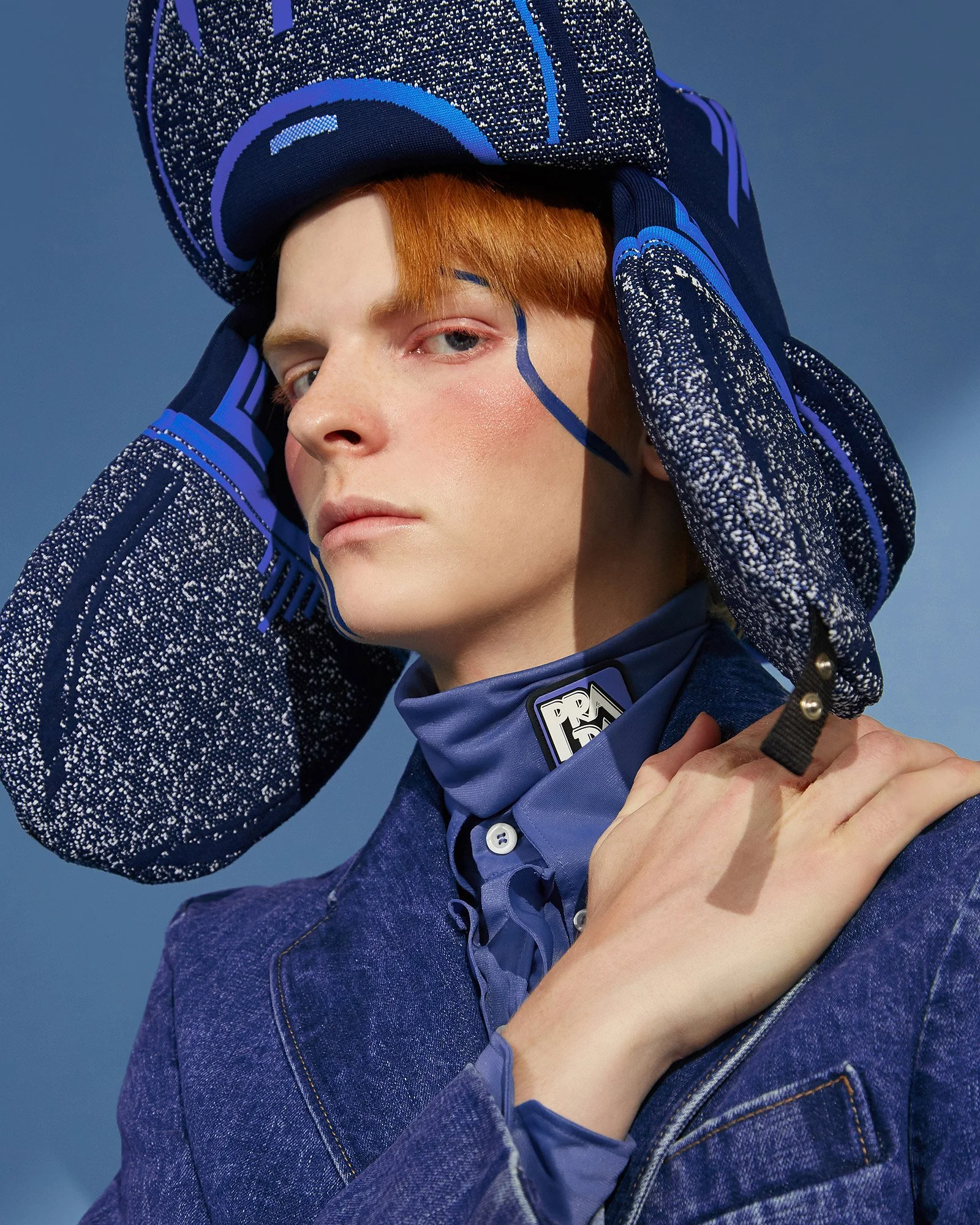 A close-up of a young person with red hair wearing a large navy and blue patterned hat, a blue jacket, and a blue scarf with the Prada logo, posing with one hand on their shoulder against a blue background.