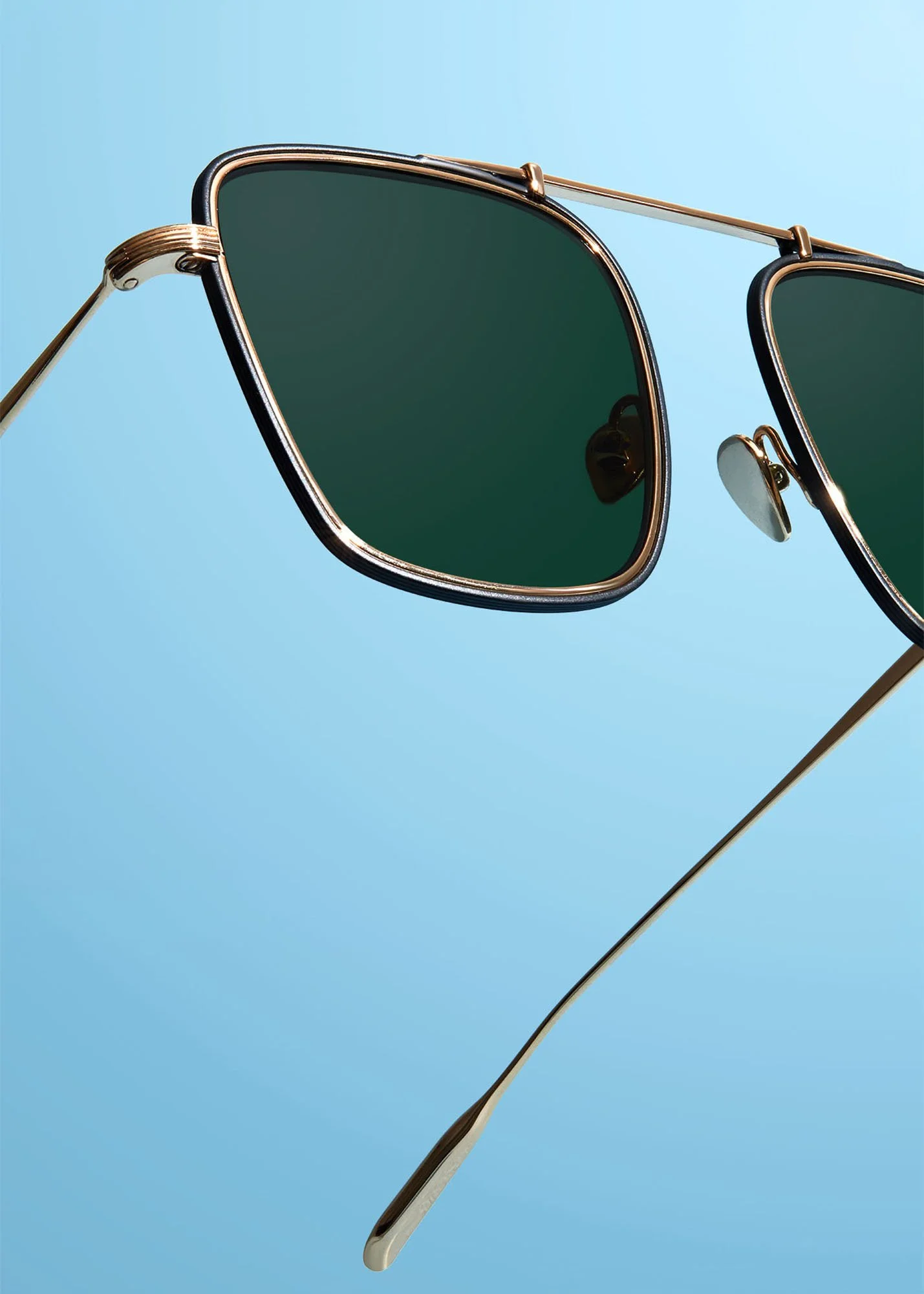 Close-up of stylish sunglasses with dark green lenses, gold-colored metal frame, and adjustable nose pads, set against a light blue background.