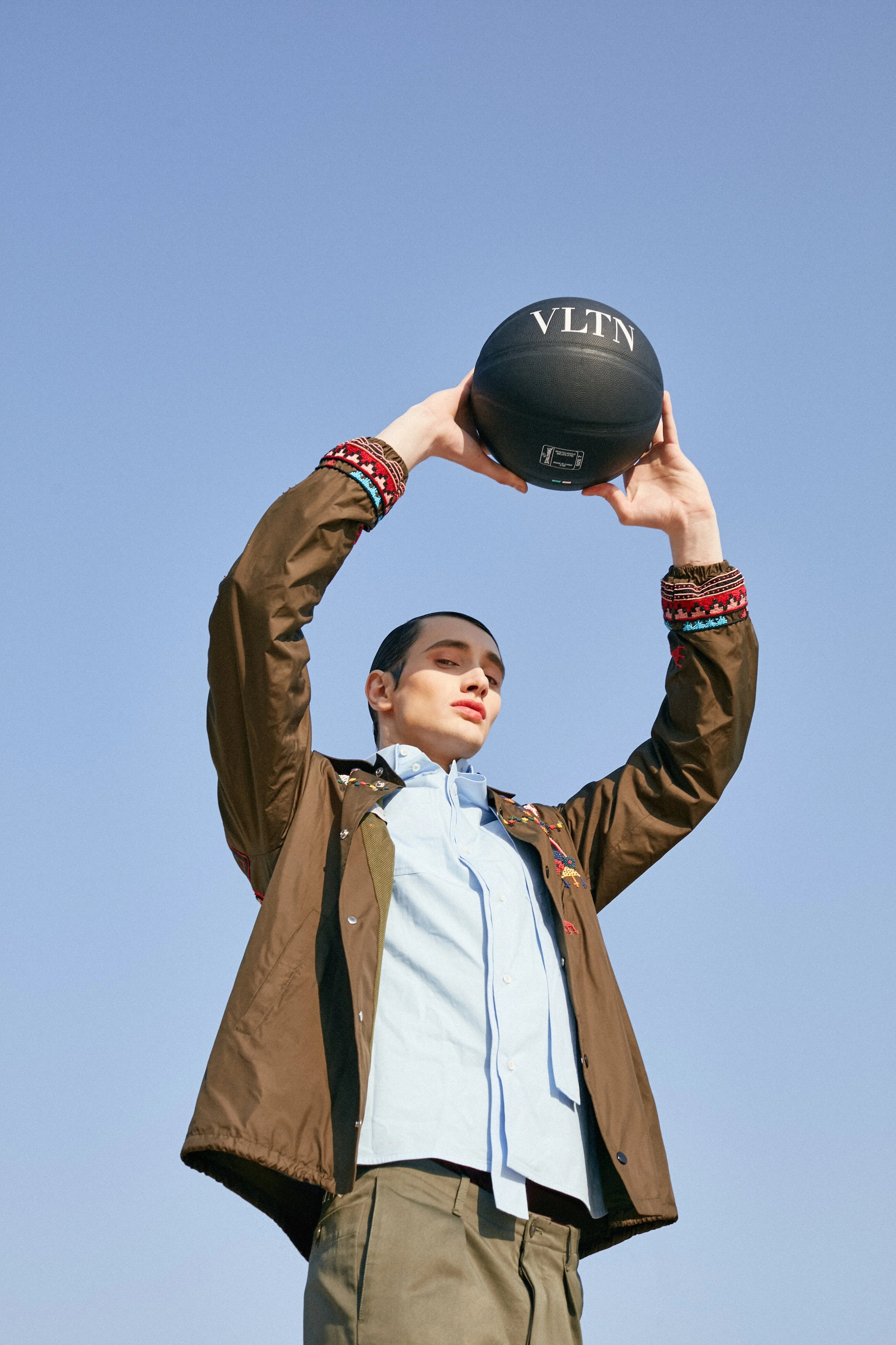 A person holding a black basketball above their head outdoors against a clear blue sky.