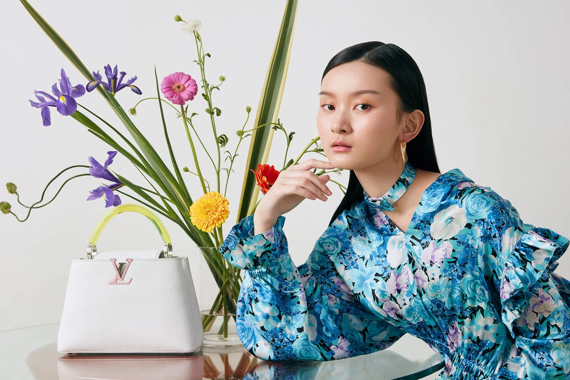 A woman wearing a blue floral dress and gold earrings is sitting next to a white designer handbag with a pink 'V' logo, surrounded by a colorful bouquet of flowers in a clear vase against a plain white background.