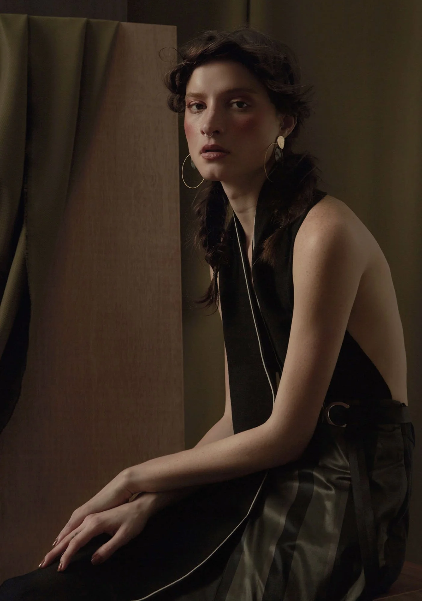 A young woman with short, curly dark hair and piercings, wearing large hoop earrings, a black sleeveless top, and striped pants, sitting in front of a wooden partition with curtains, looking at the camera in a dimly lit room.