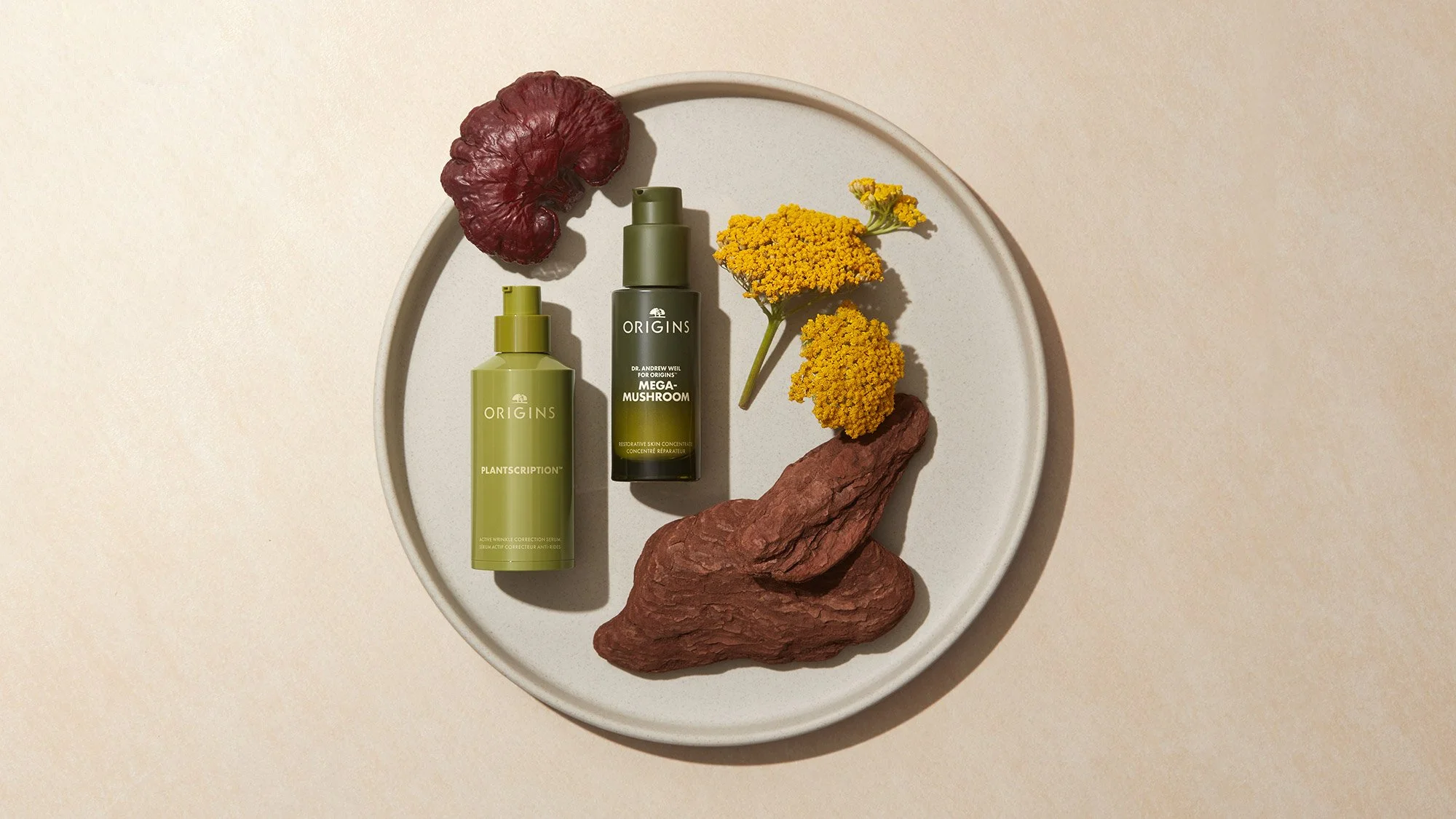 Skincare products on a round white plate, including two green bottles labeled Origins, with yellow flowers and dried red plant pieces around them.