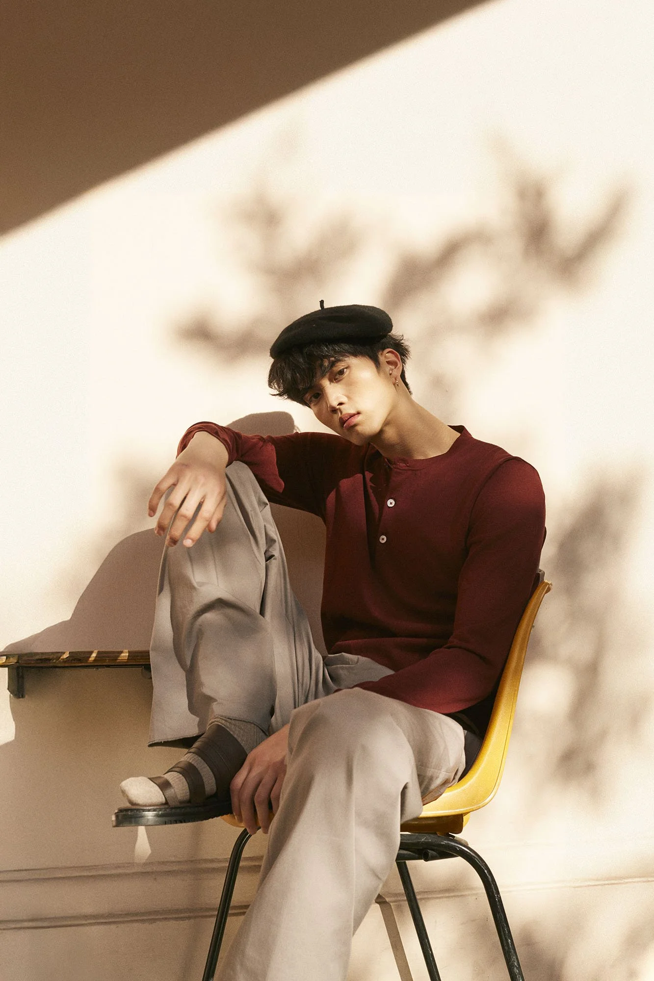 A young man with dark hair and earrings wearing a black beret, red long-sleeve shirt, and beige pants, sitting on a yellow chair against a plain wall with a shadowy plant silhouette. He has one leg crossed over the other, with a contemplative express