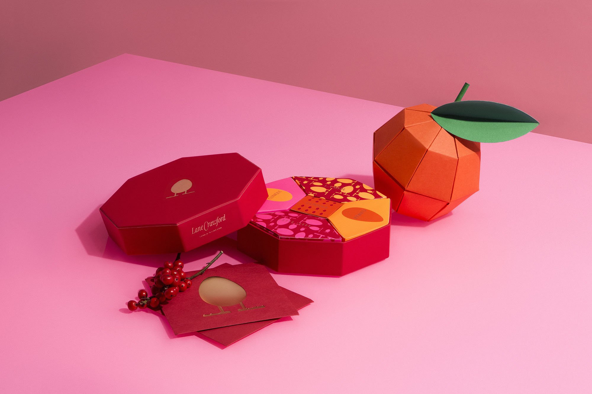 Stacked paper orange with green leaves, red and pink gift boxes with fruit-themed designs, and red berries on pink surface with pink background.