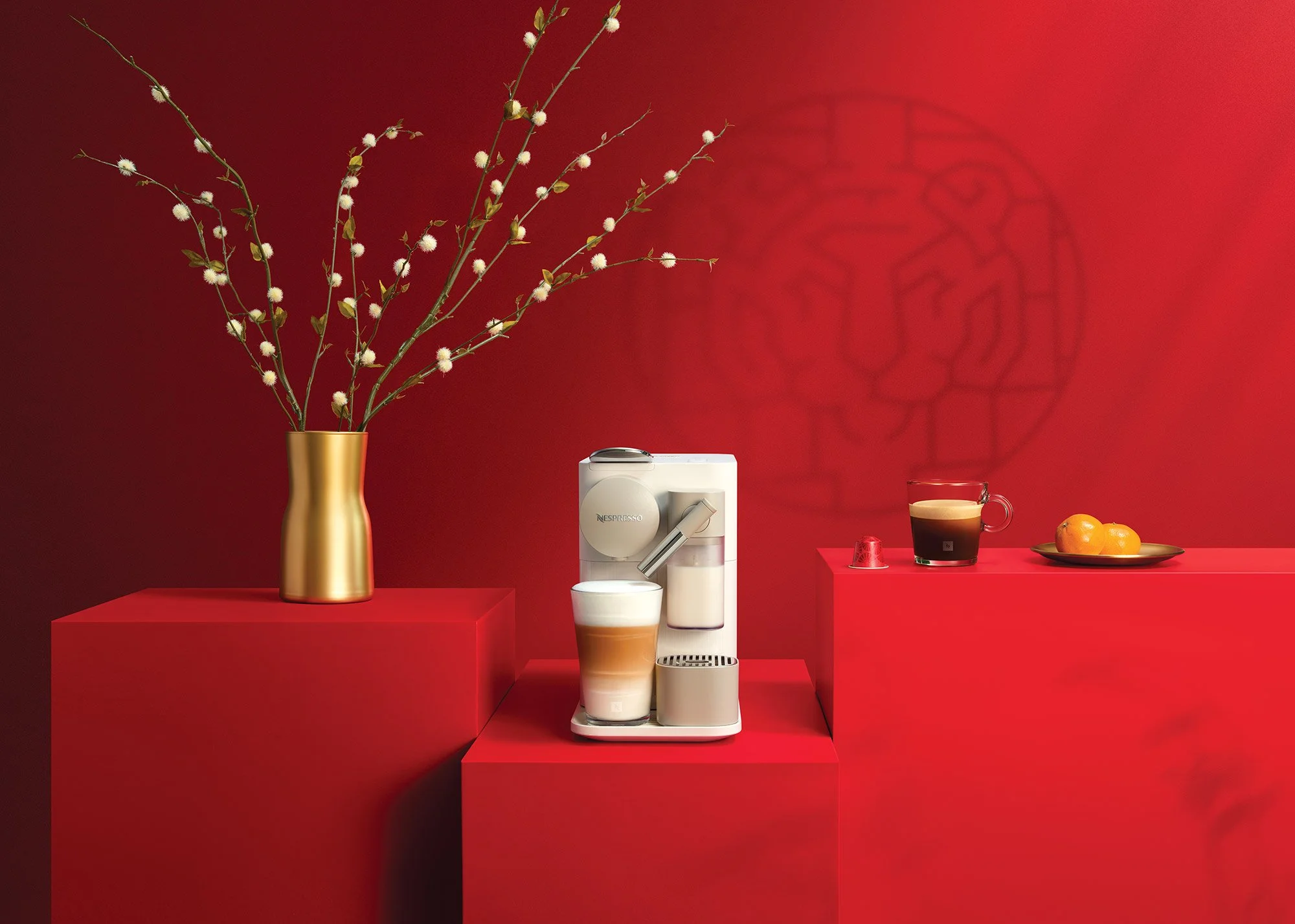 Red background with a gold vase holding flowering branches, a white Nespresso coffee machine with a cup, and a plate with oranges on red platforms.