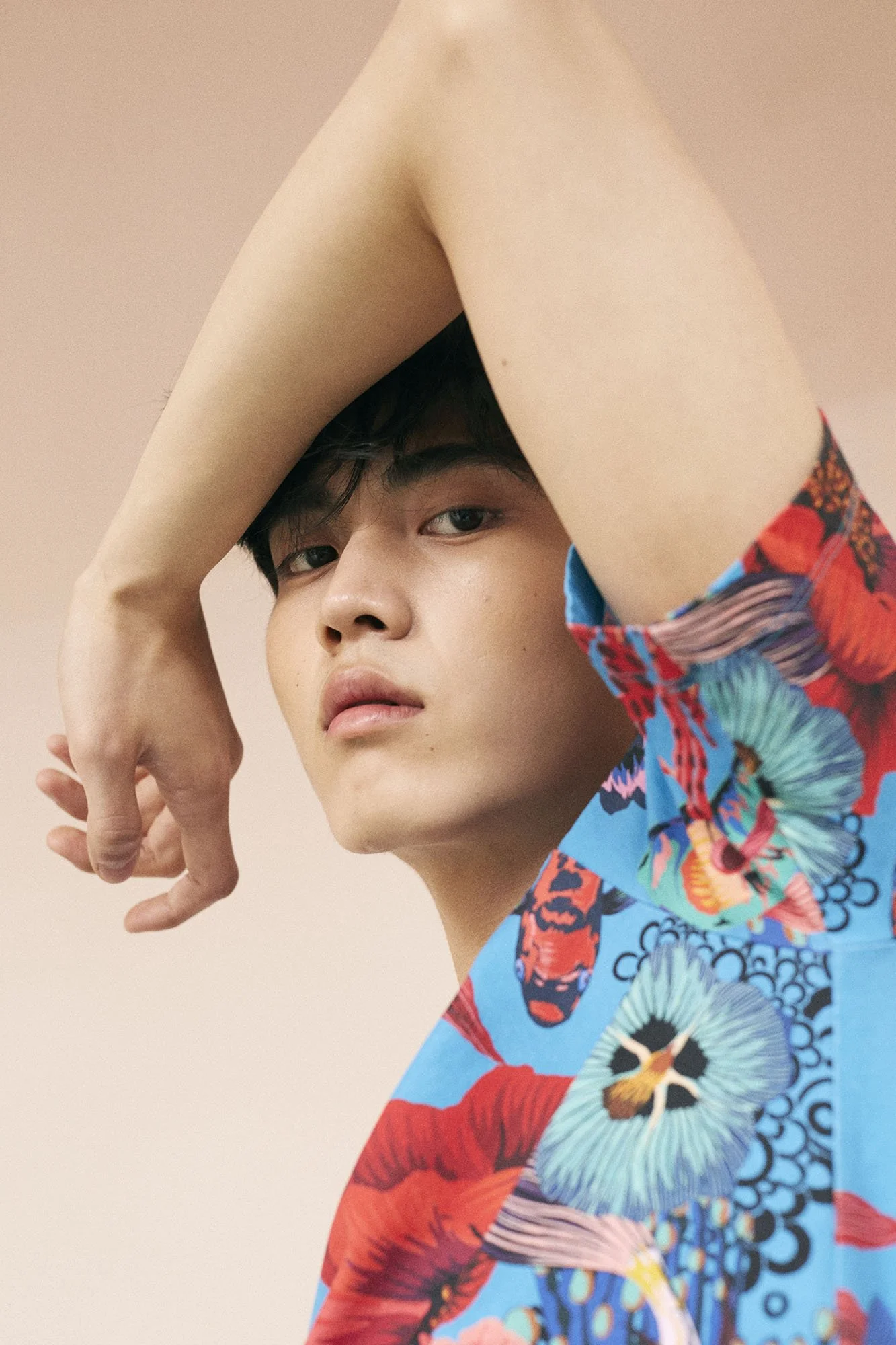 A young man with fair skin and dark hair, wearing a brightly colored shirt with a tropical floral pattern, posing with one arm raised above his head against a plain, light-colored background.