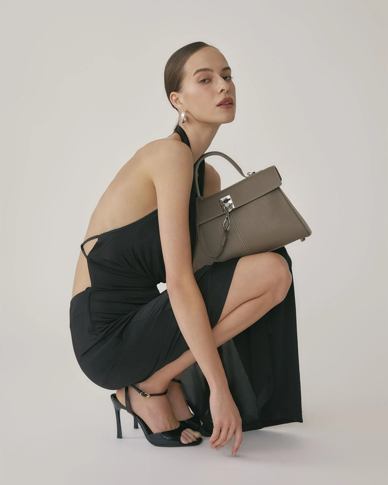 A woman in black heels and a black dress is crouching with her knees bent and holding a taupe handbag. She has light skin, slicked-back hair, silver earrings, and a confident expression, set against a plain light background.