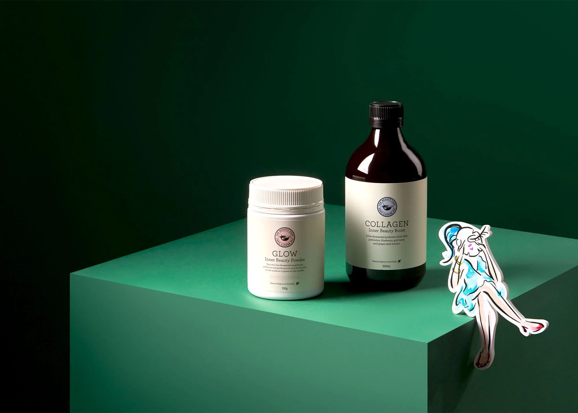 Beauty products including a white jar labeled "GLOW Inner Beauty Powder" and a dark bottle labeled "COLLAGEN Inner Beauty Boost" displayed on a green platform alongside a cartoon sticker of a unicorn with blue and pink mane.