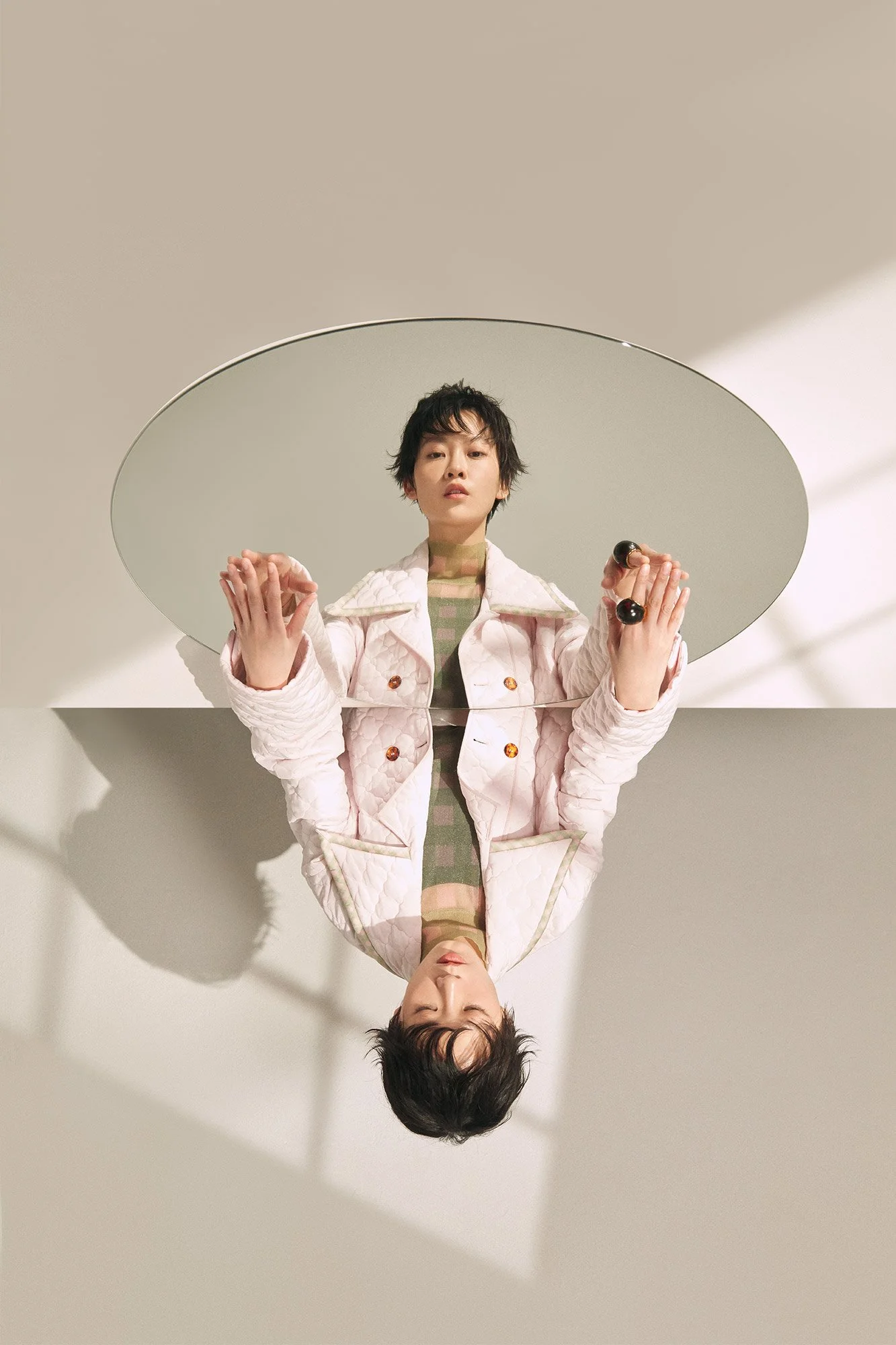 A person standing in front of a mirror, reflected on a round mirror on a light-colored wall. The person has short, dark hair and is wearing a light pink quilted jacket with large buttons over a beige and green checkered shirt.