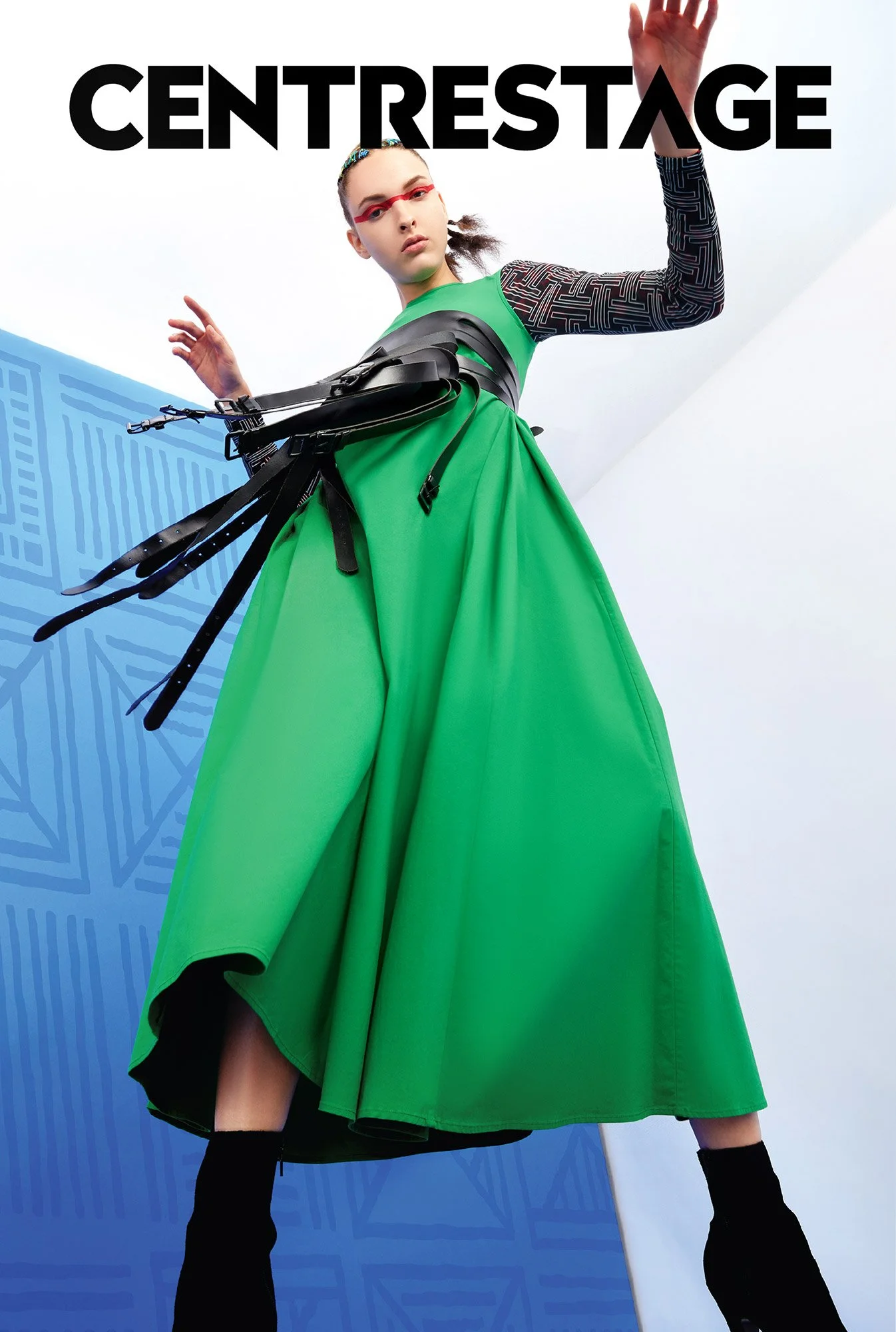 Fashion model wearing a green dress with black accessories and geometric patterned sleeves, standing against a white and blue background with THEATRE logo.