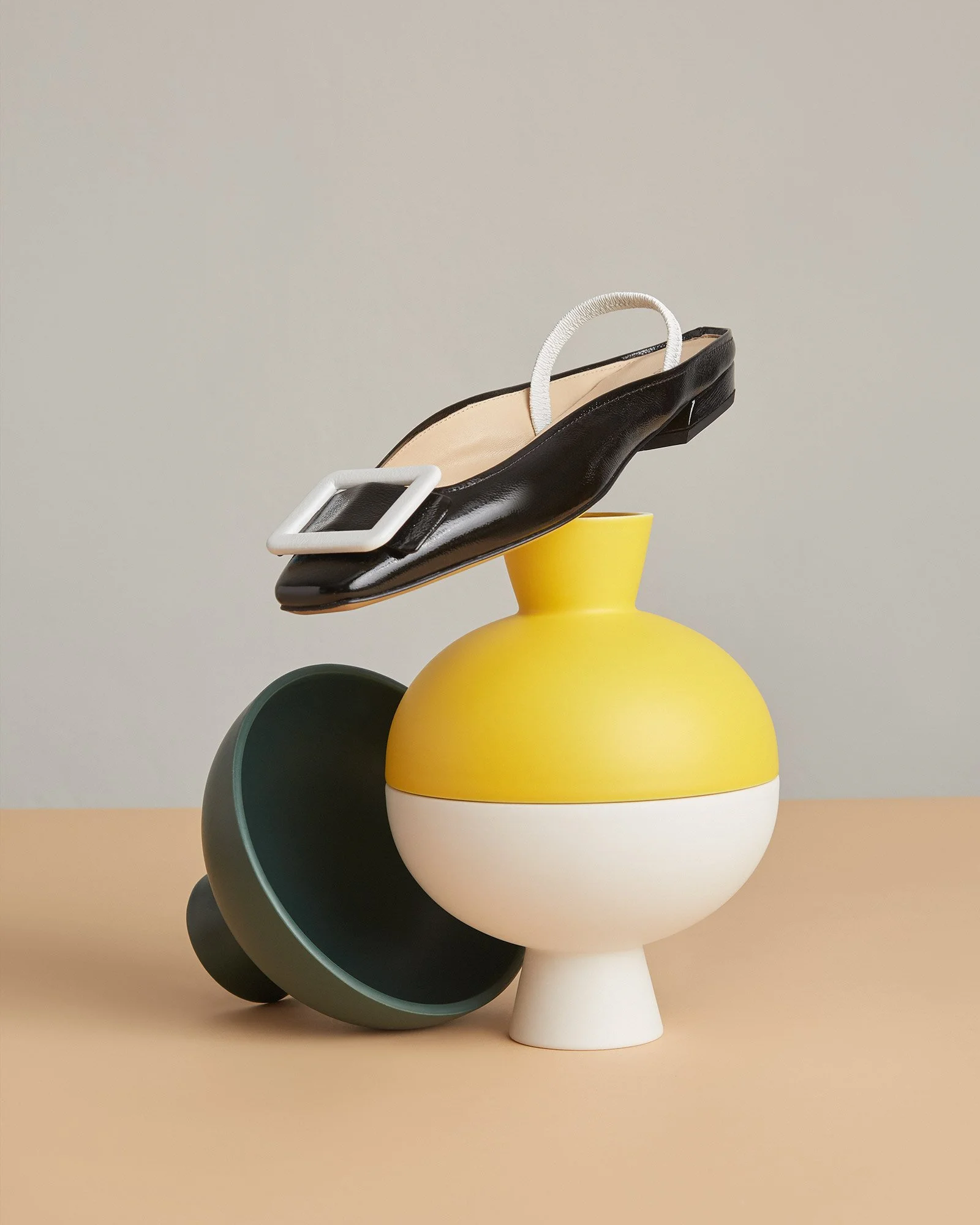 A black patent leather women's shoe with a square buckle, positioned atop a colorful abstract ceramic vase with a yellow top, white middle, and beige bottom, on a beige surface with a neutral background.