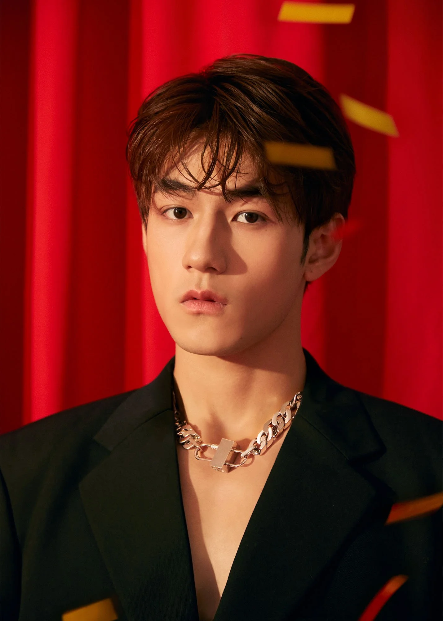 A young man with dark, styled hair, wearing a black blazer and a chunky gold chain necklace, standing in front of red curtains.