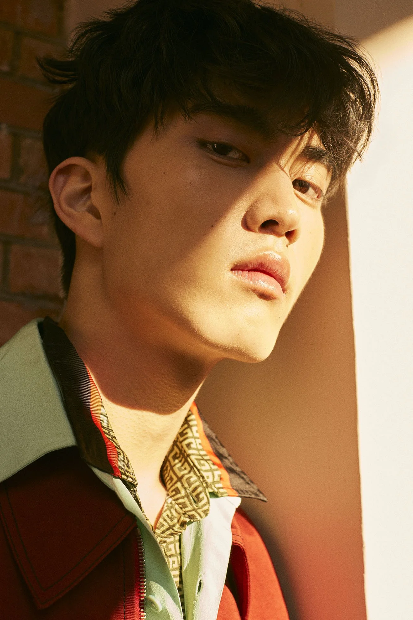 Close-up of a young man with dark hair, wearing a patterned shirt and a red and green jacket, looking slightly to the side with sunlight illuminating his face.