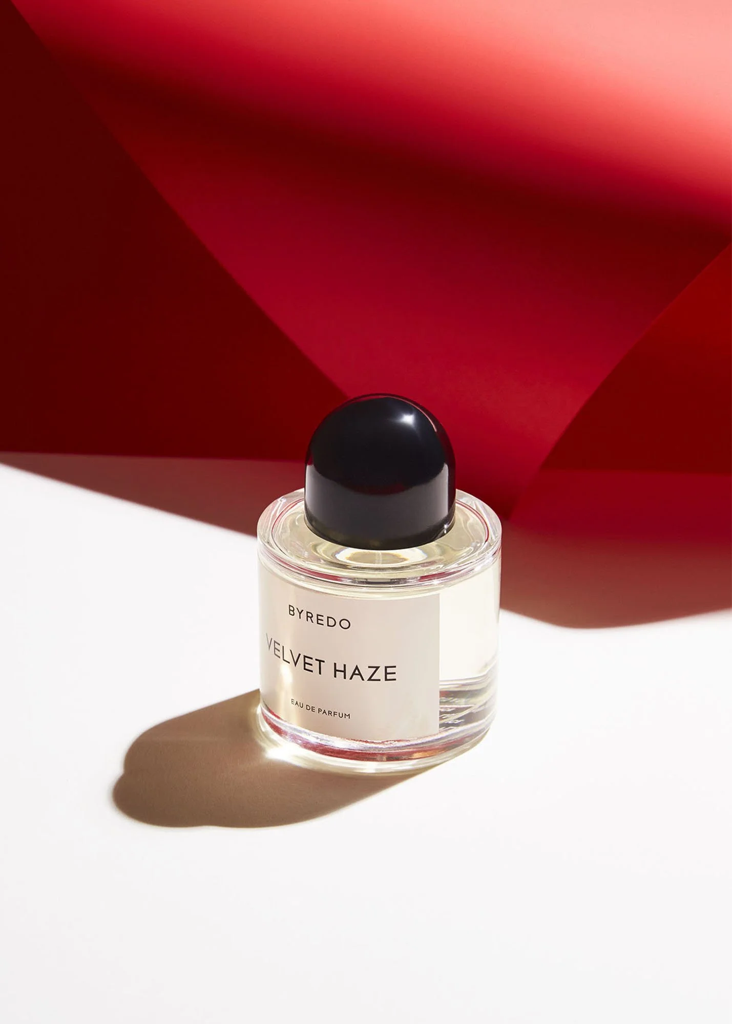 A perfume bottle labeled 'Velvet Haze' by Byredo on a white surface, with a shadow cast by the bottle. A red background with a curved shadow adds a contrasting backdrop.