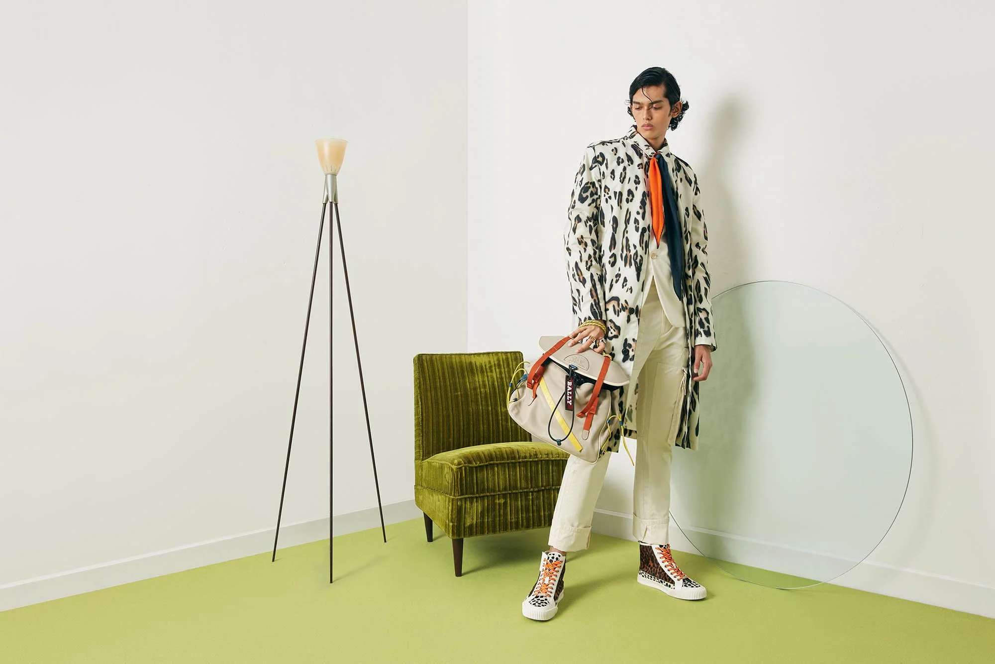Person standing in a minimalist room holding a beige bag with orange straps, wearing animal print coat, cream pants, and leopard print sneakers, with a green velvet chair, tripod lamp, and a clear circular glass object in the background.