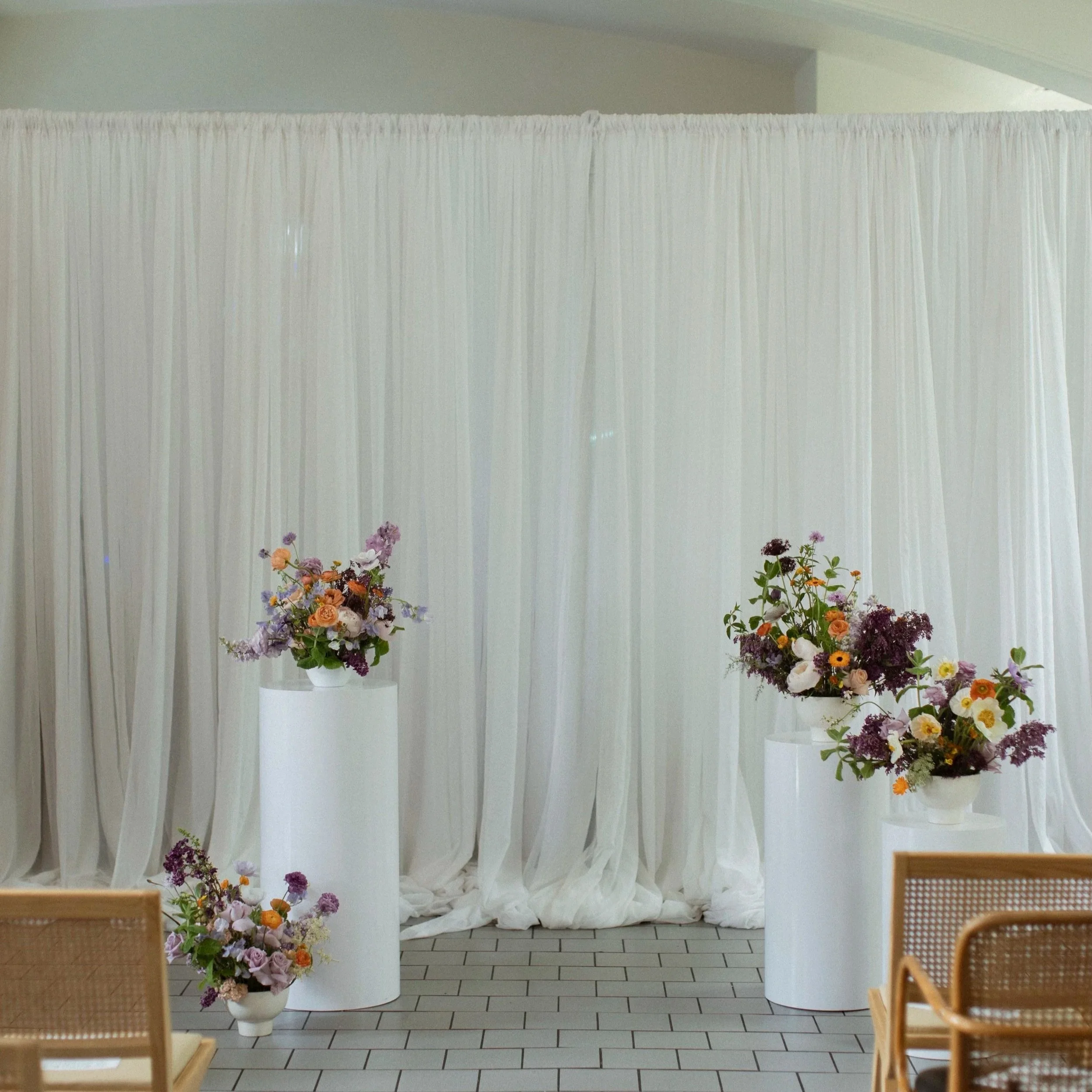 Decorative floral arrangements in white vases placed on white cylindrical pedestals in front of a white draped fabric backdrop, with chairs in the foreground.