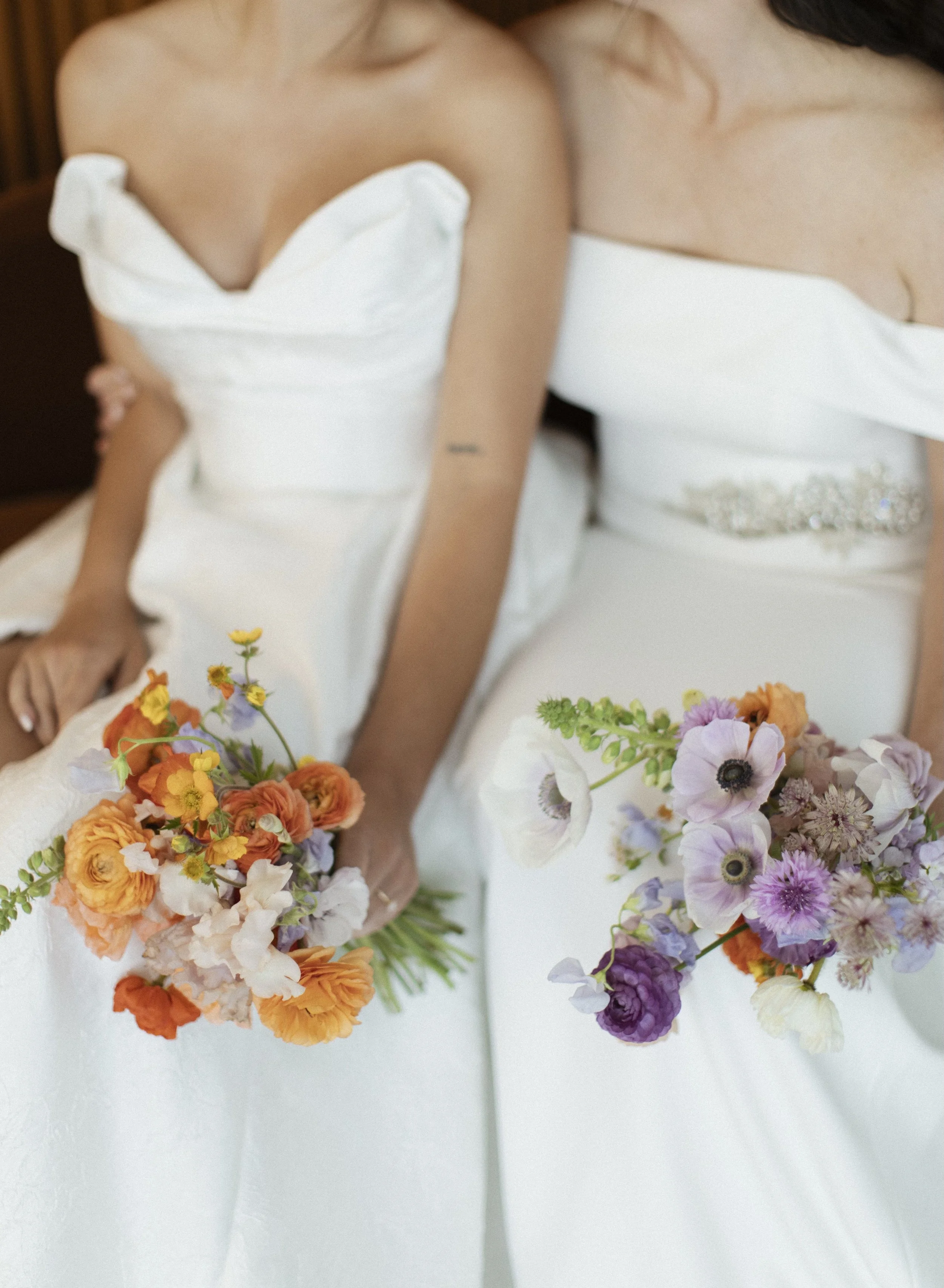 Two women in white dresses holding colorful flower bouquets, sitting together.