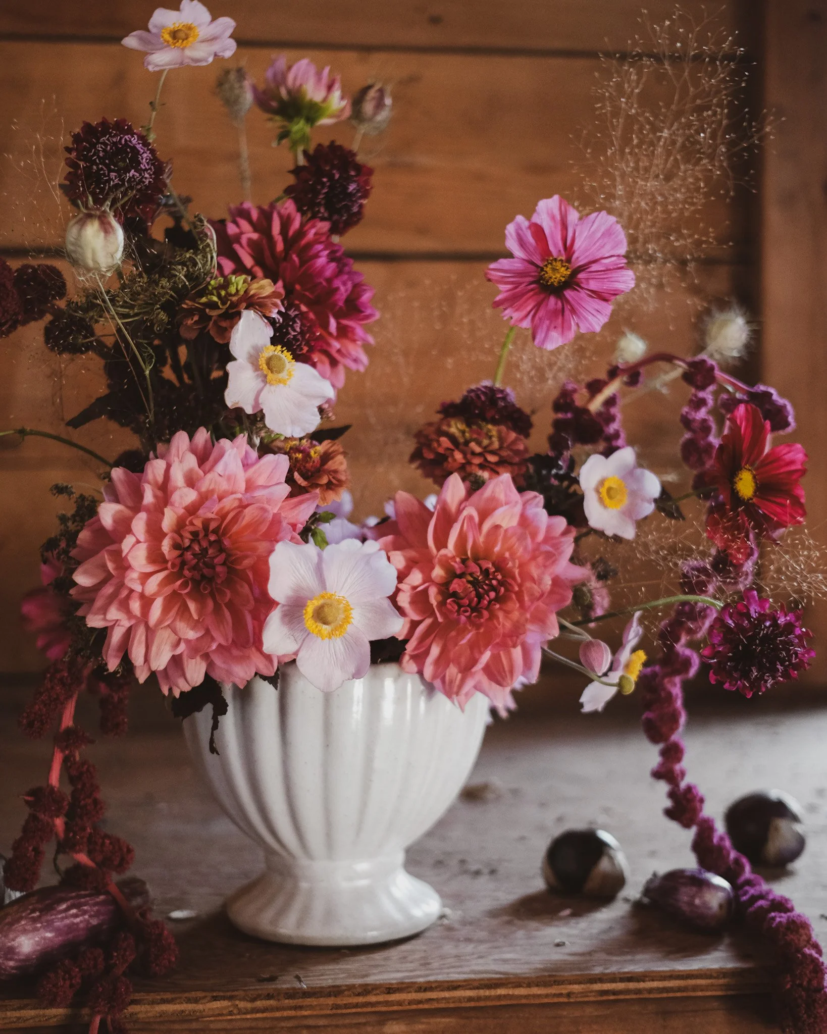 A white ceramic vase filled with pink, white, and purple flowers, including dahlias and cosmos, on a wooden surface with a wooden wall background.