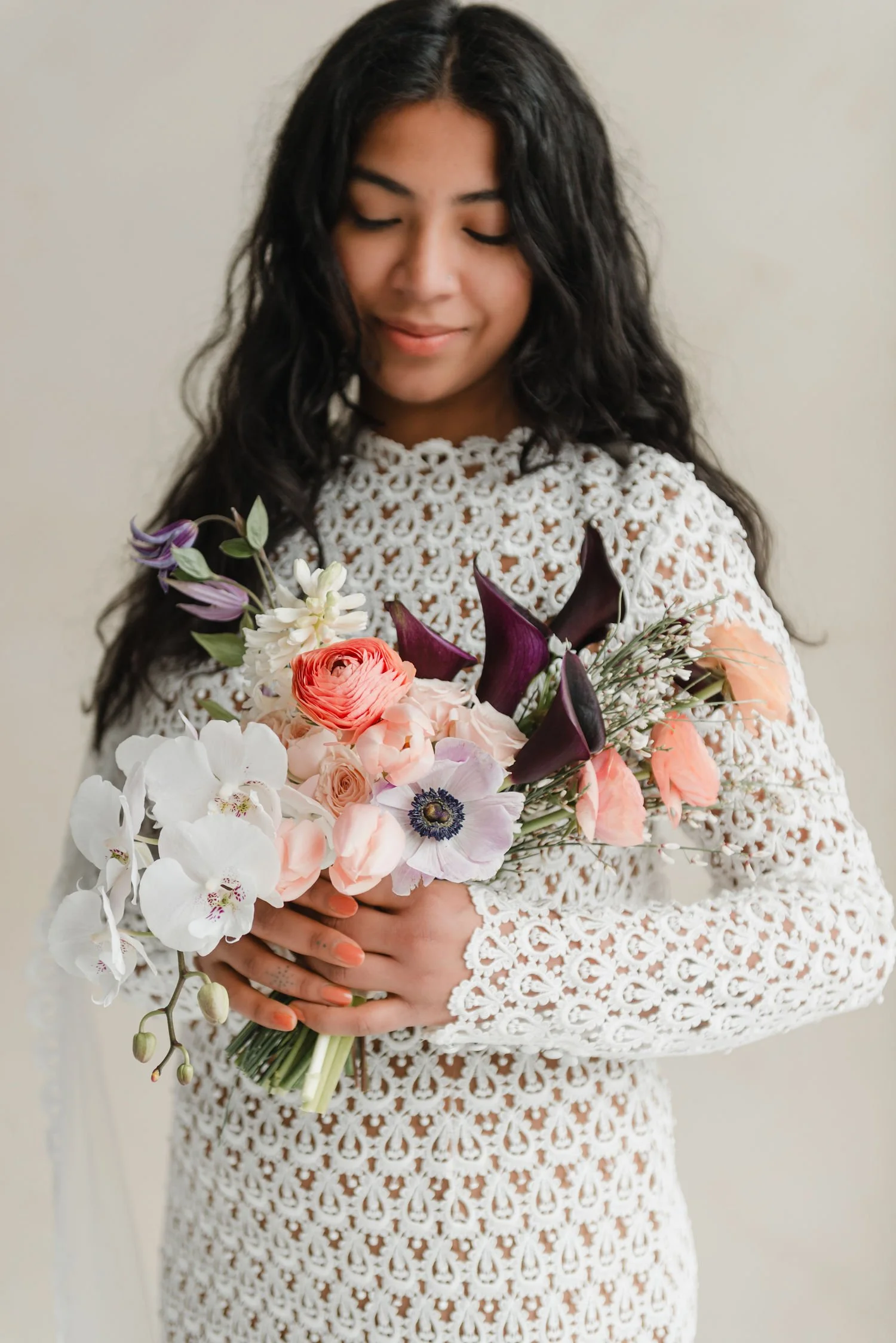 A woman with dark, wavy hair and closed eyes holds a colorful bouquet of flowers, wearing a white lace dress.
