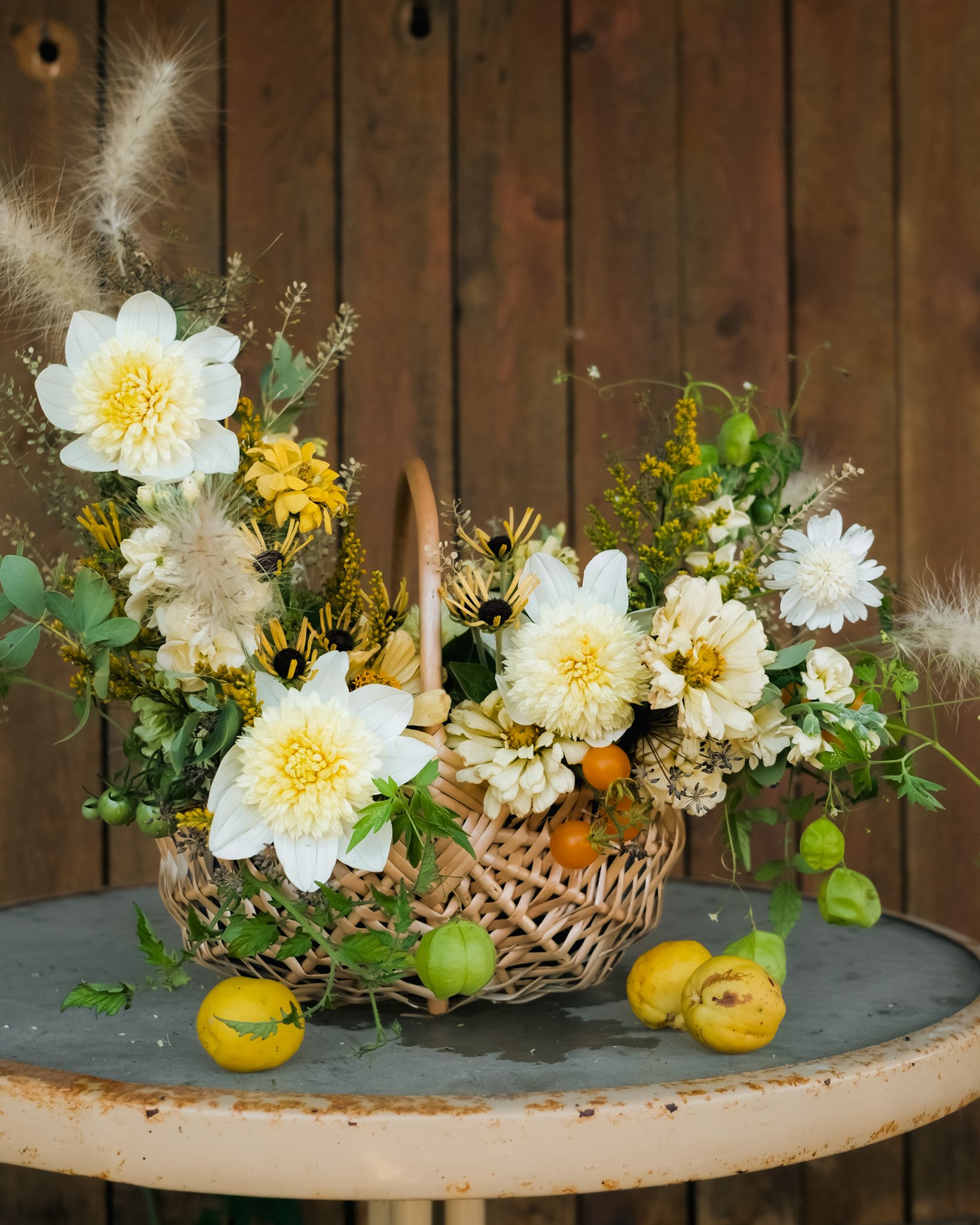 A woven basket filled with white and yellow flowers, green foliage, and small green and orange tomatoes, set on a rustic round table in front of a wooden wall.