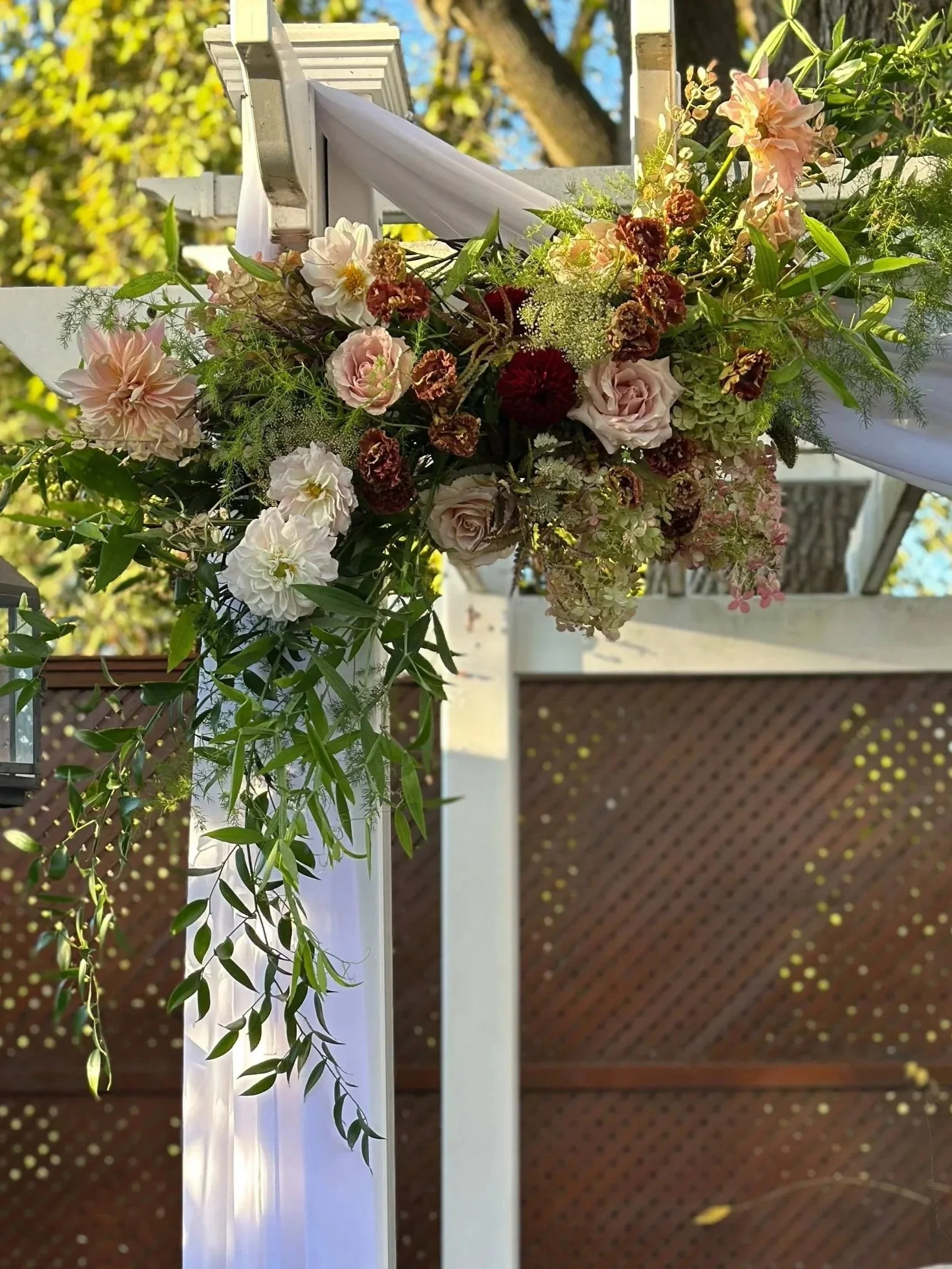 Decorative floral arrangement with pink, white, and burgundy flowers, along with greenery, attached to a white outdoor structure, with a wooden lattice fence in the background.